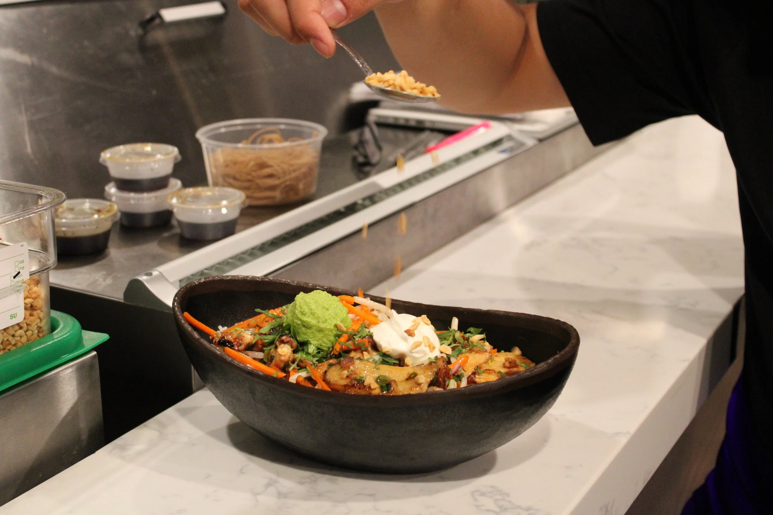 Person adding toppings to a bowl of food on a kitchen counter, with various ingredients and sauces visible in the background.
