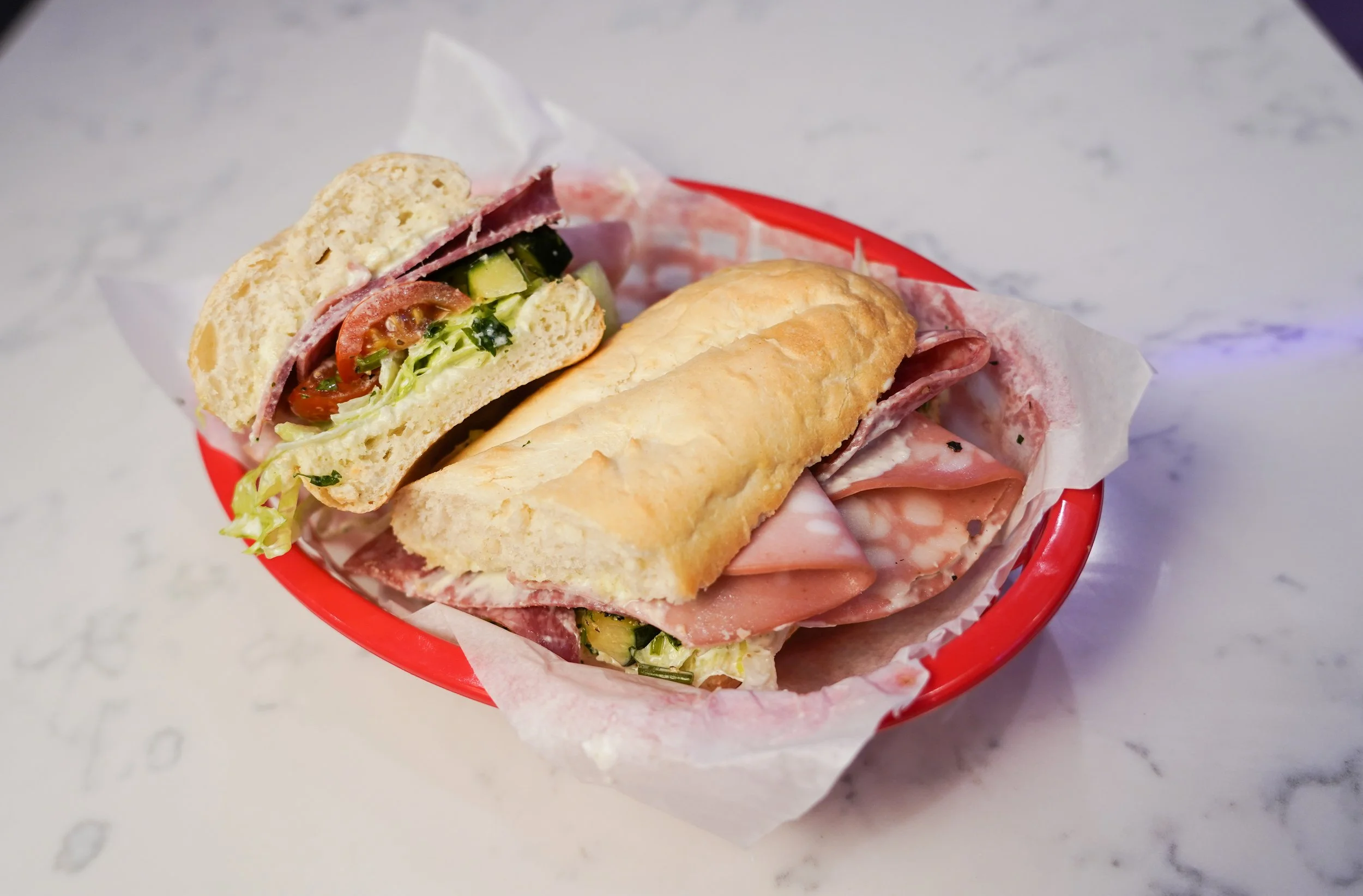 Sandwich with meats, lettuce, tomato, and cucumber in a red basket on a table.