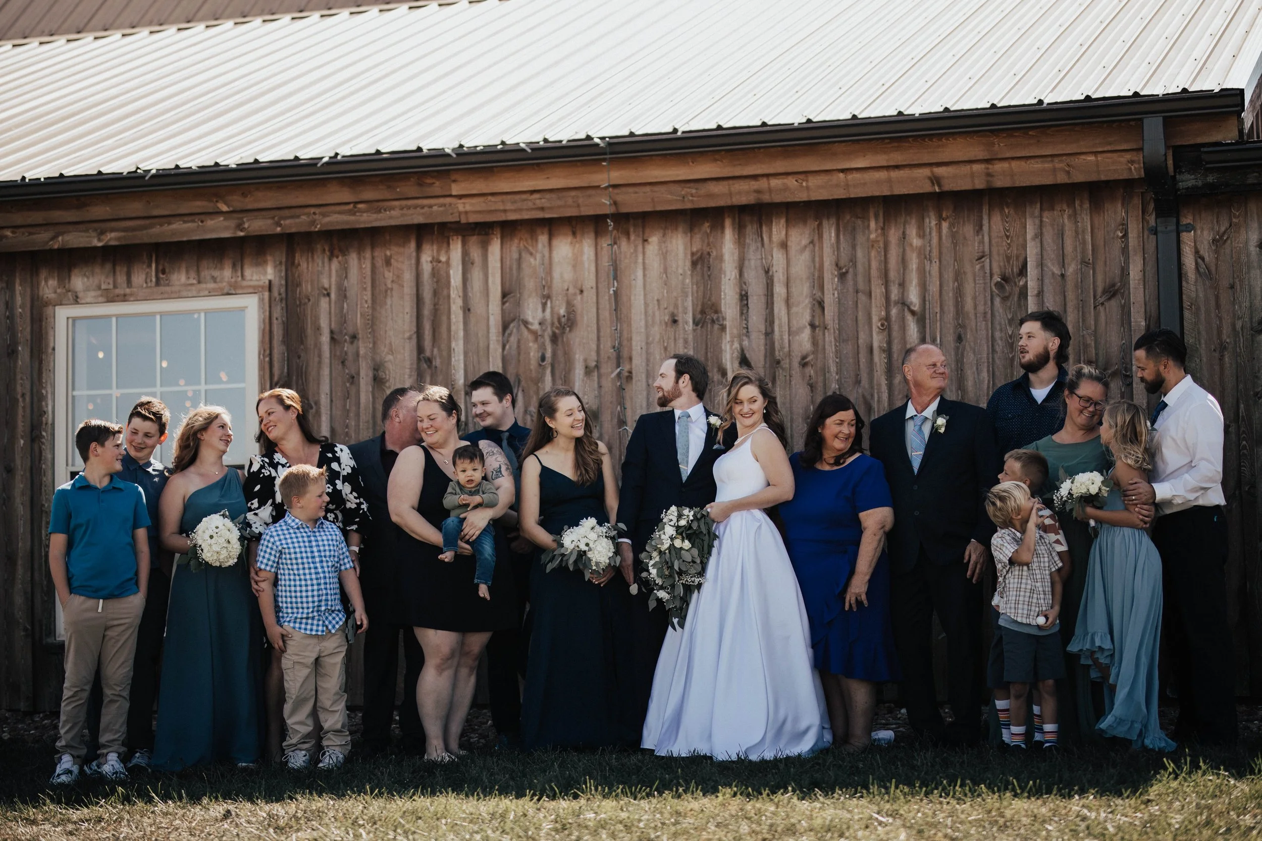 A large group of people, including men, women, and children, dressed in formal attire, standing outdoors in front of a wooden building with a metal roof, celebrating a wedding.