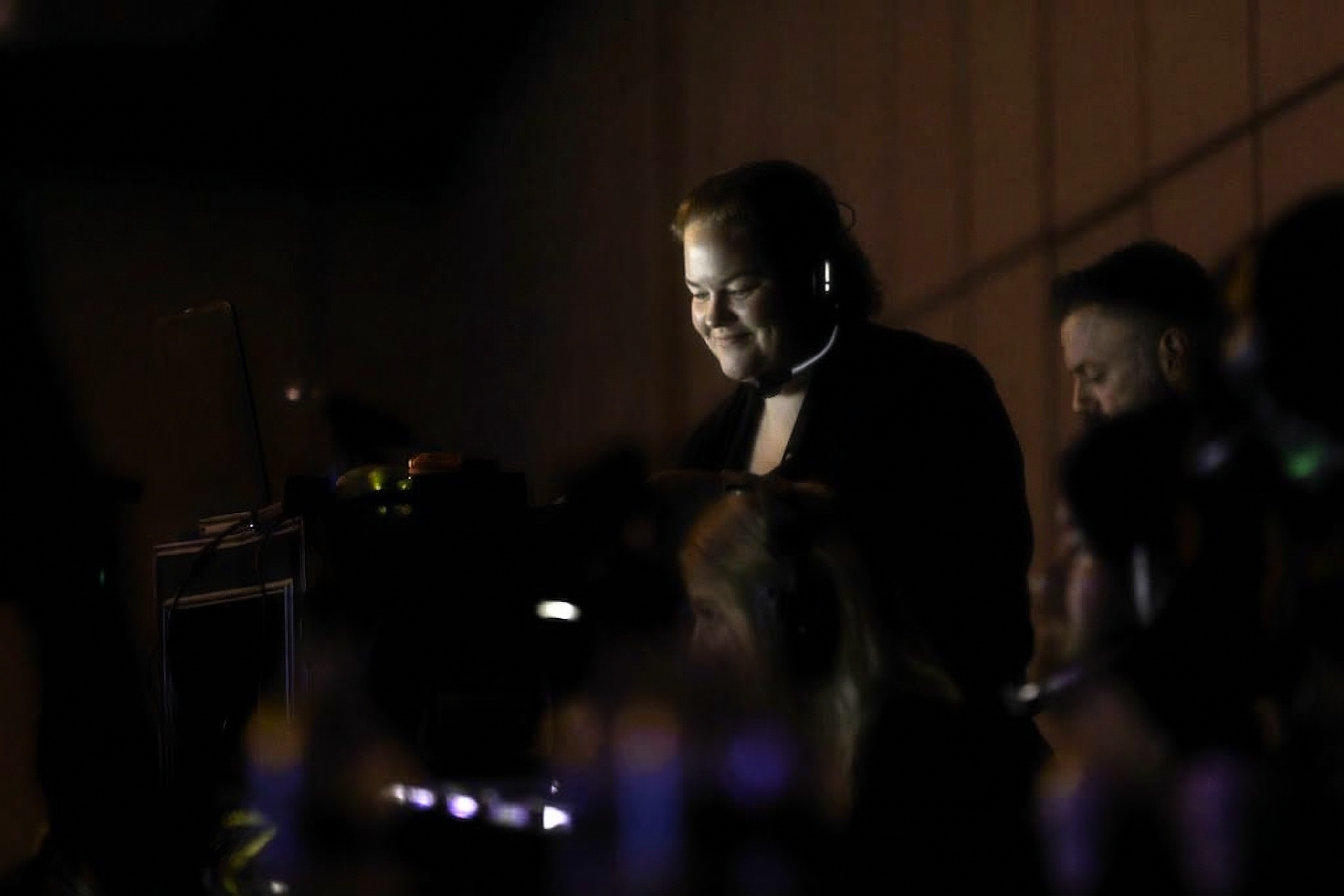 A woman in a control room wearing a headset, surrounded by dark equipment and other people, smiling and working on a computer.