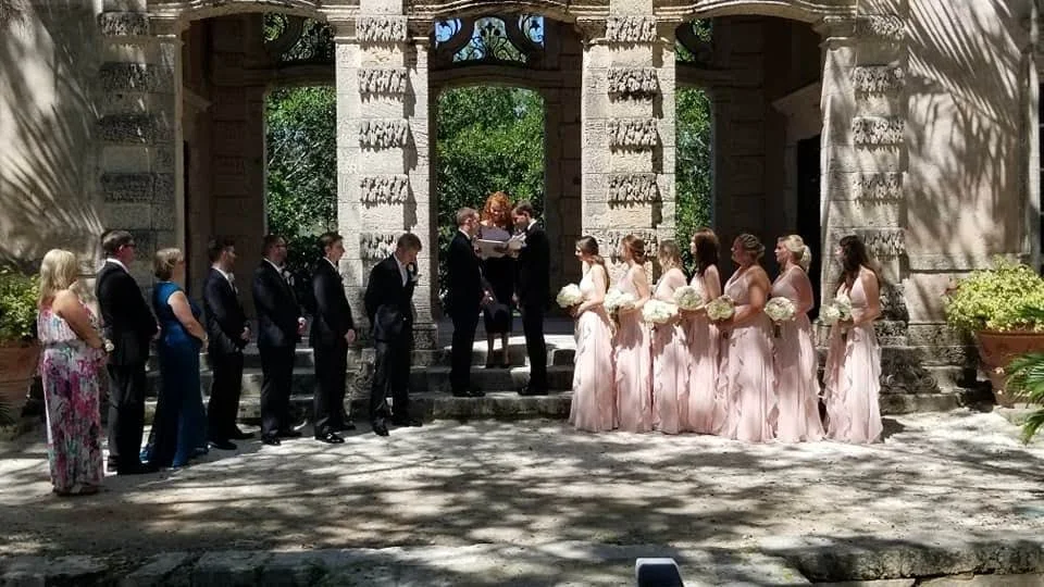 A wedding ceremony taking place outdoors with the bride and groom exchanging vows, surrounded by bridesmaids holding bouquets and groomsmen dressed in suits.