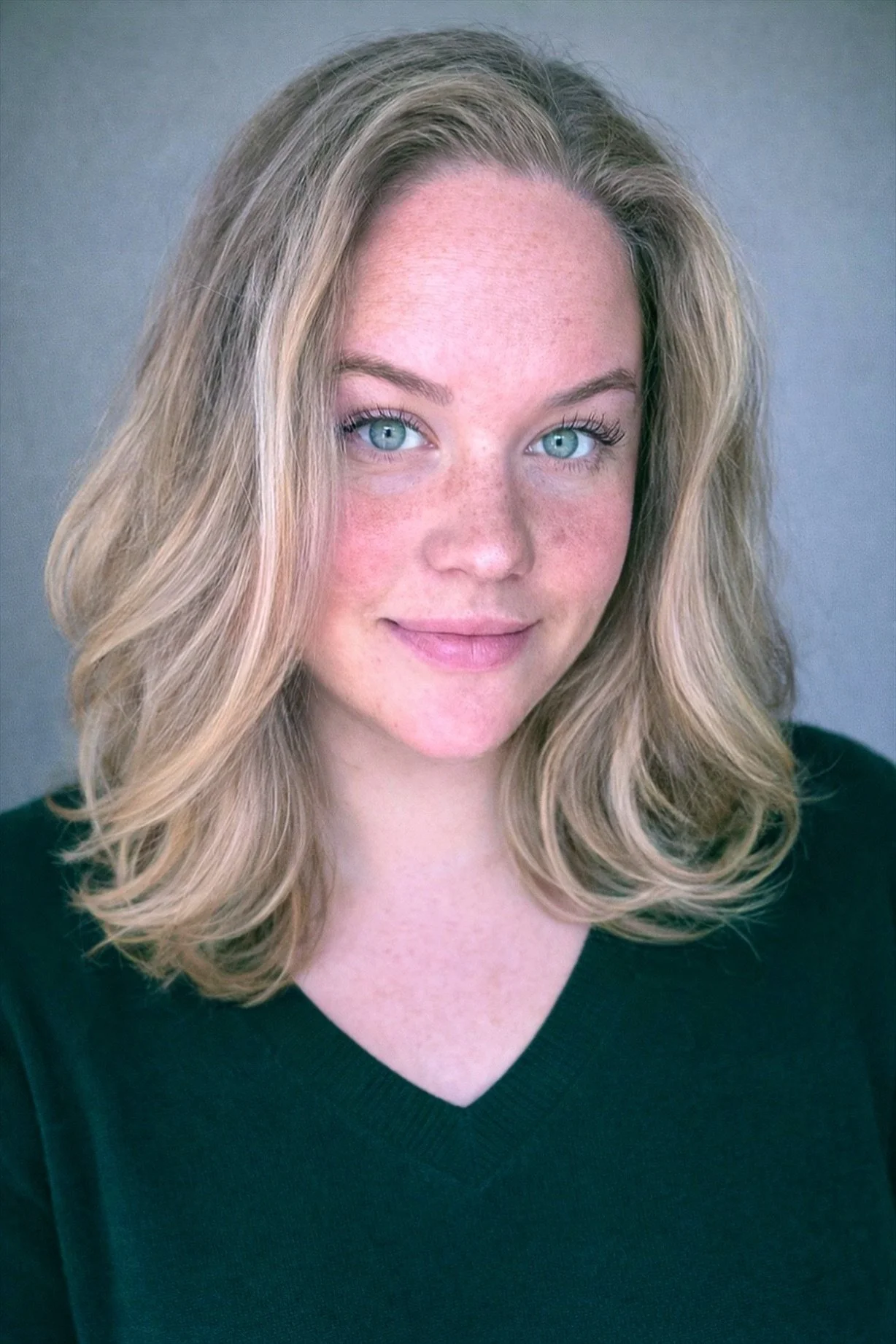 Close-up portrait of a young woman with blonde hair, blue eyes, and light skin, wearing a dark green V-neck top and smiling slightly, against a gray background.