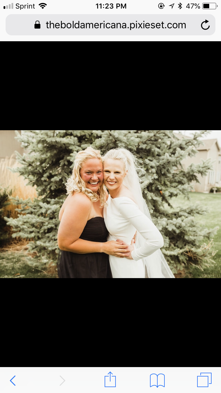 Two women smiling and hugging outdoors in front of a pine tree, one wearing a black strapless dress and the other in a white wedding dress with a veil, during a wedding celebration.