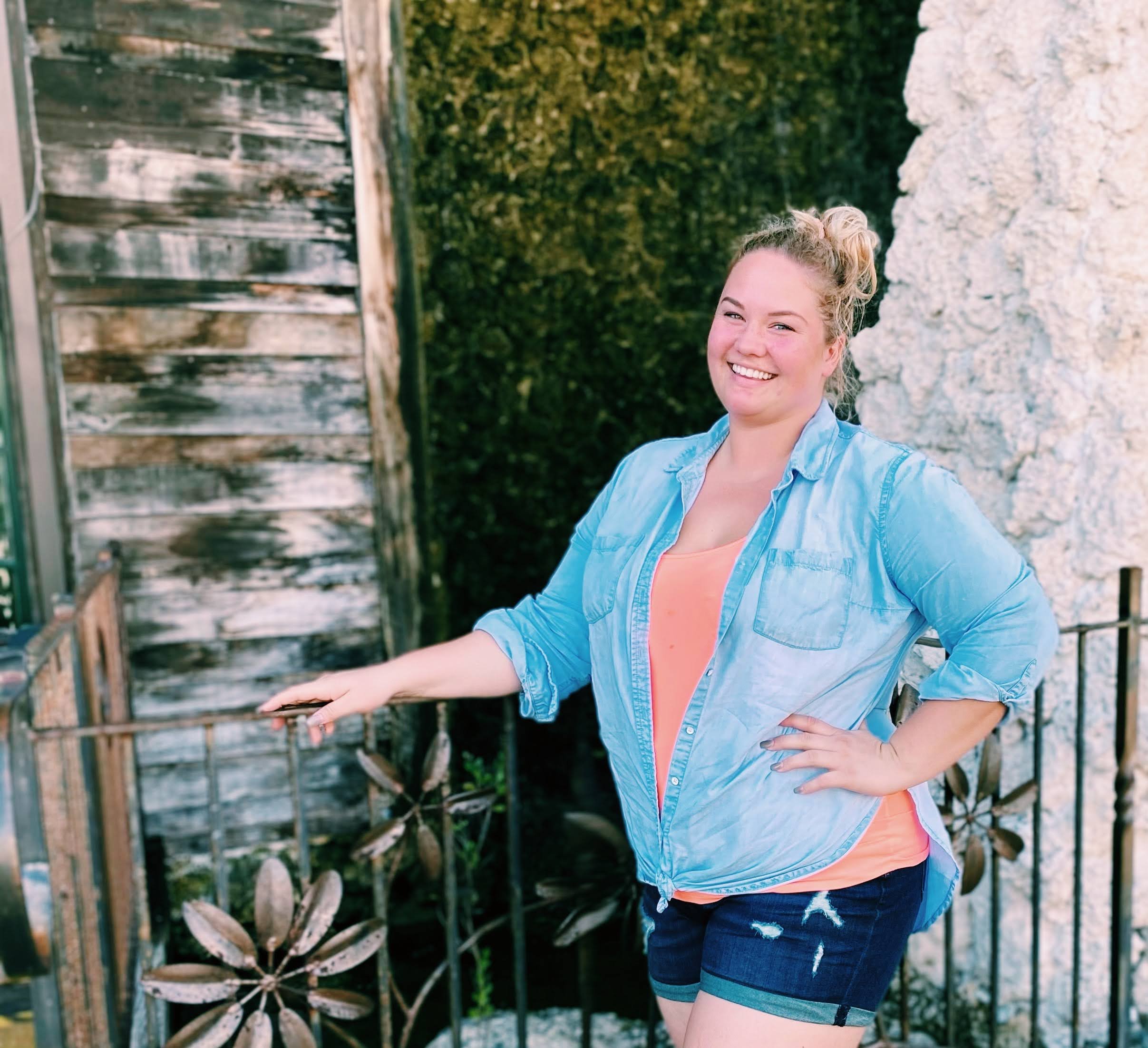 A young woman with blonde hair in a bun, smiling and posing outdoors. She is wearing a light blue denim shirt over an orange top and dark denim shorts, standing next to a metal fence with a flower design, with greenery and an old wooden wall in the background.