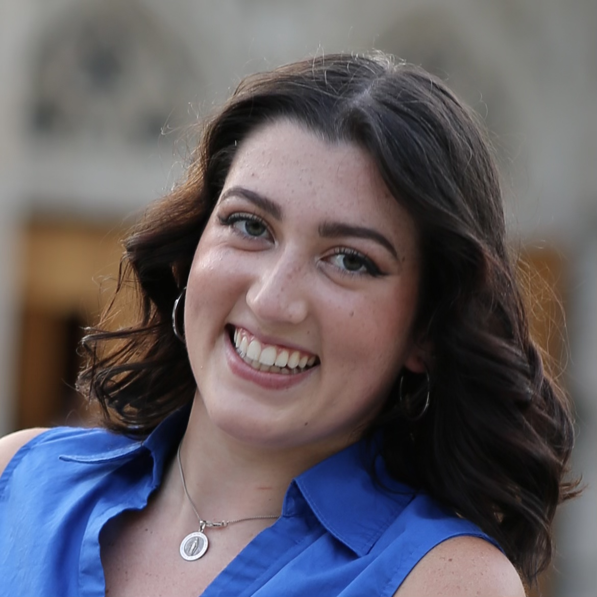 Smiling person with wavy hair wearing a blue shirt and necklace.