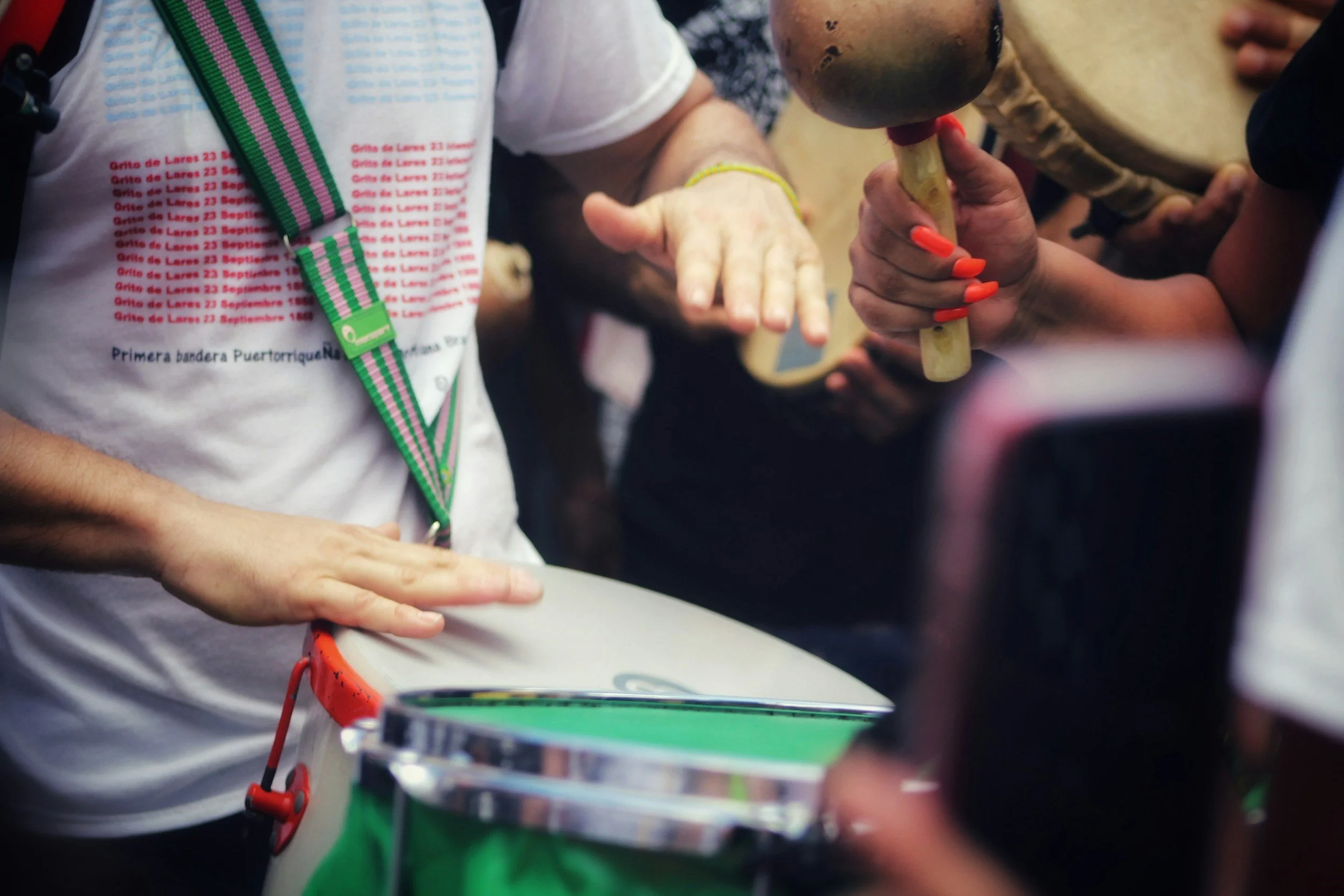 Closeup of a person playing a drum while people play shakers, bongos, and other percussion instruments in the background.