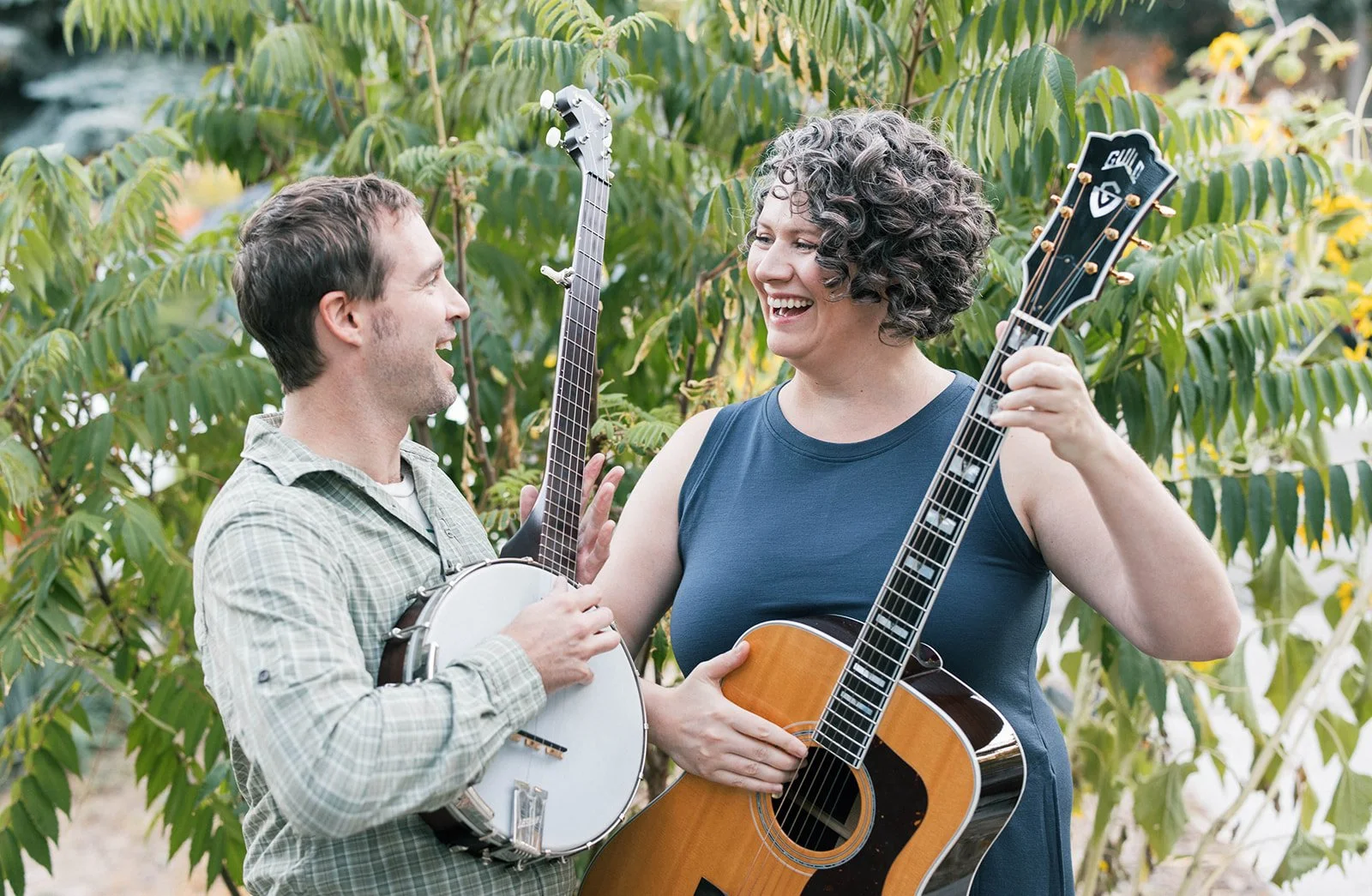 Campfire Music Collective teachers Angie and Marcus Marianthi smiling outside at their outdoor music studio. Angie holds a guitar, and Marcus holds a banjo.