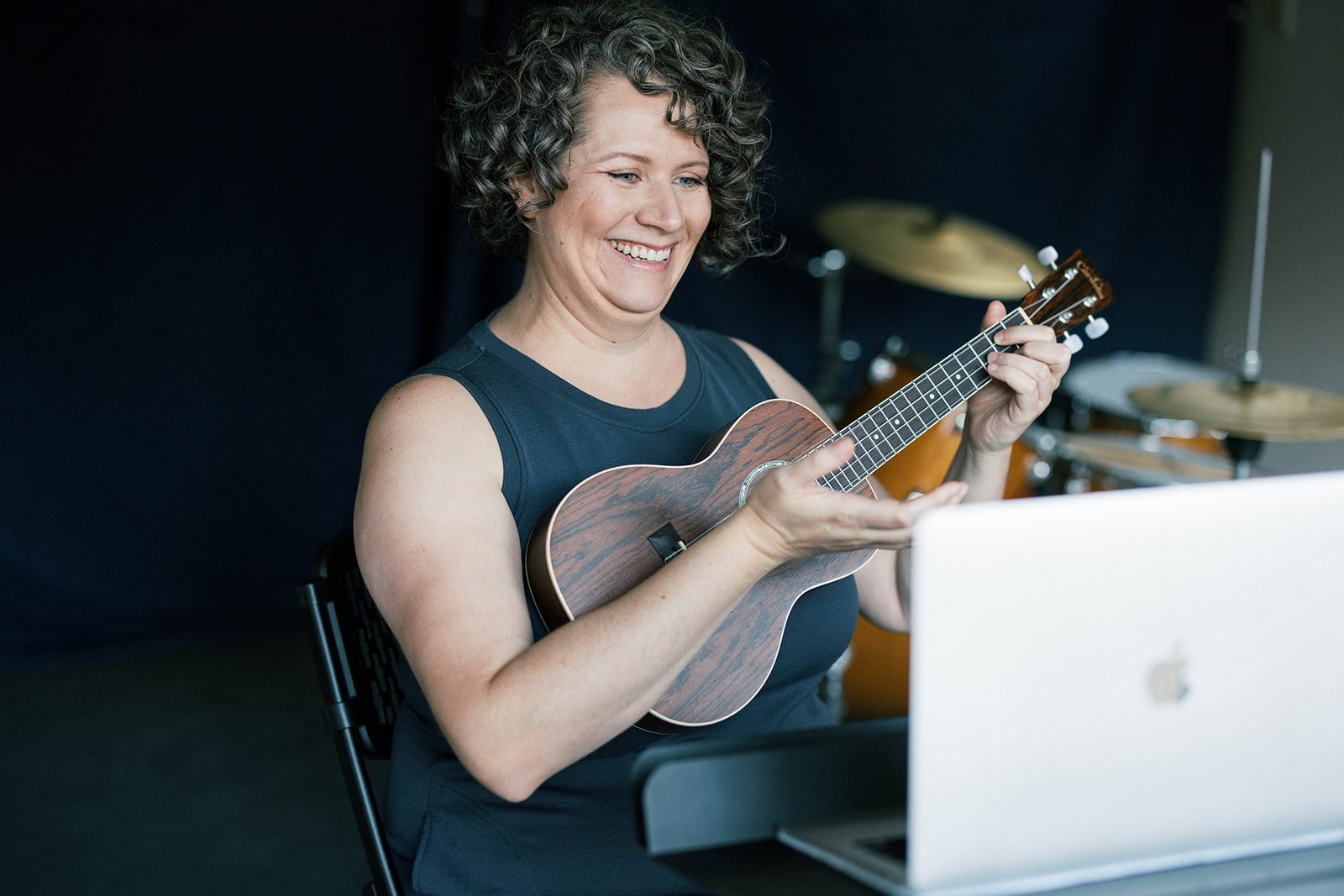 Campfire Music Collective teacher Angie Marianthi holds a ukulele while smiling and talking to students through a live online session on her computer.