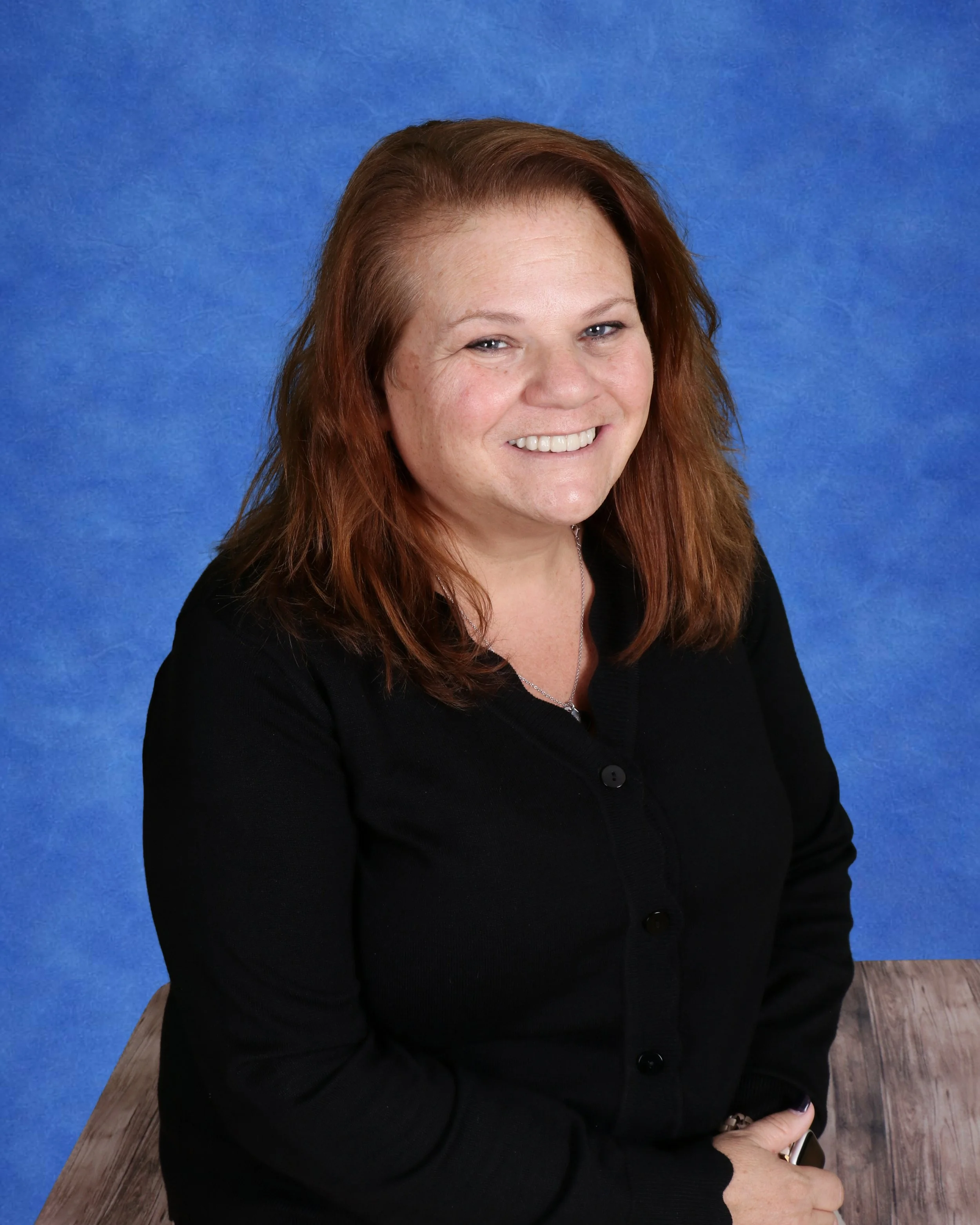 A woman with shoulder-length brown hair, smiling, wearing a dark gray dress with a black neckline and a white short-sleeve shirt underneath, standing in front of a light-colored brick wall.