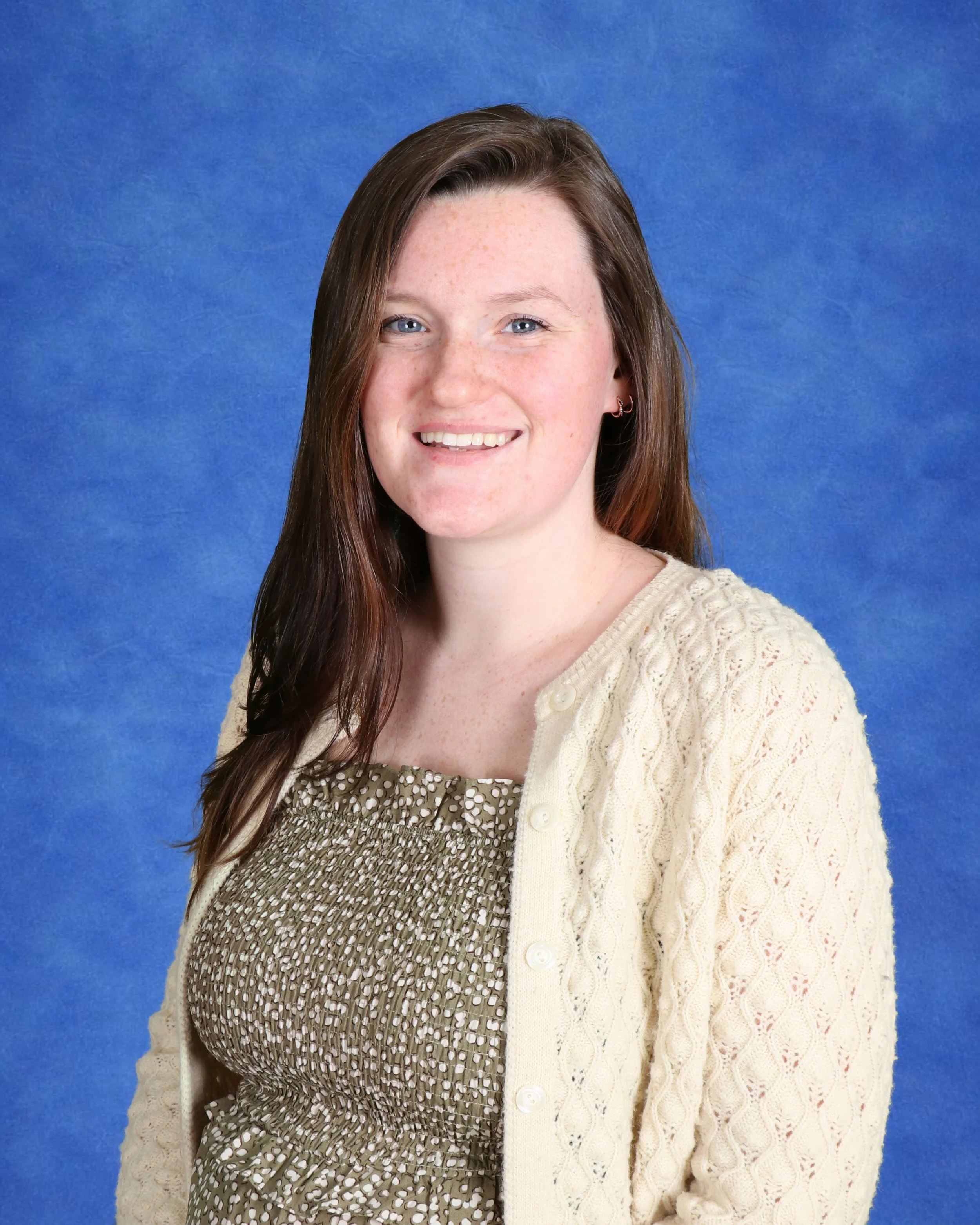 A young woman with long brown hair wearing a yellow floral top, smiling softly at the camera against a plain light background.