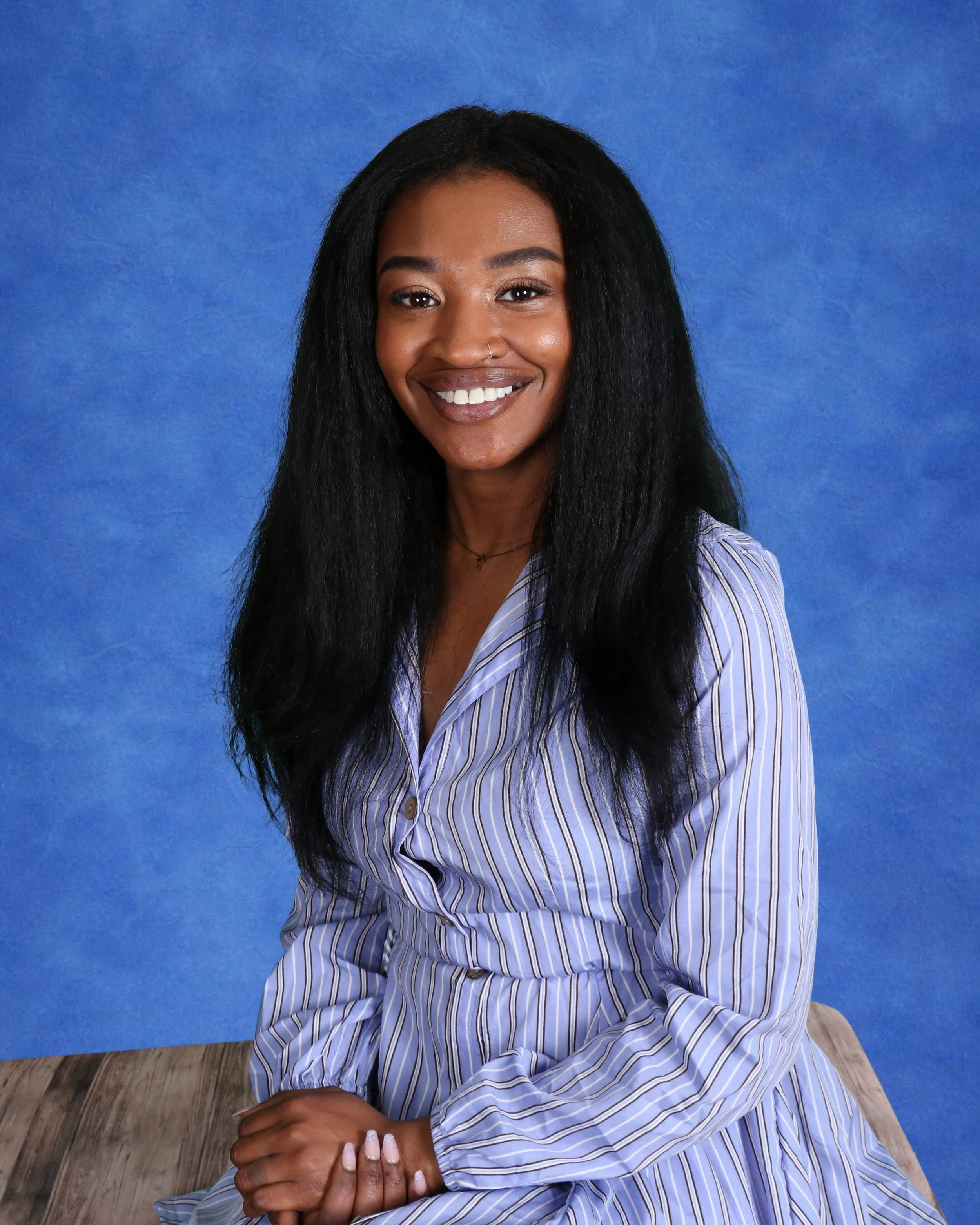 Portrait of a young woman with shoulder-length hair, smiling, wearing a black quilted vest over a black sweater, standing against a light-colored cinder block wall.