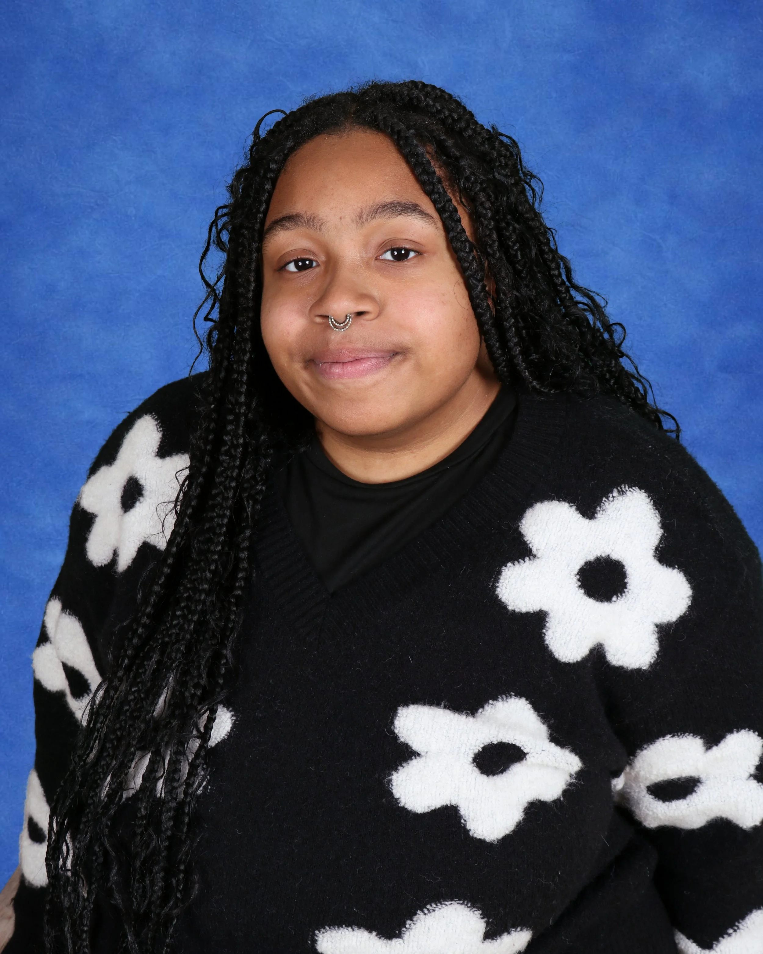 Young woman with long braids, glasses, and septum piercing standing against a light gray brick wall.