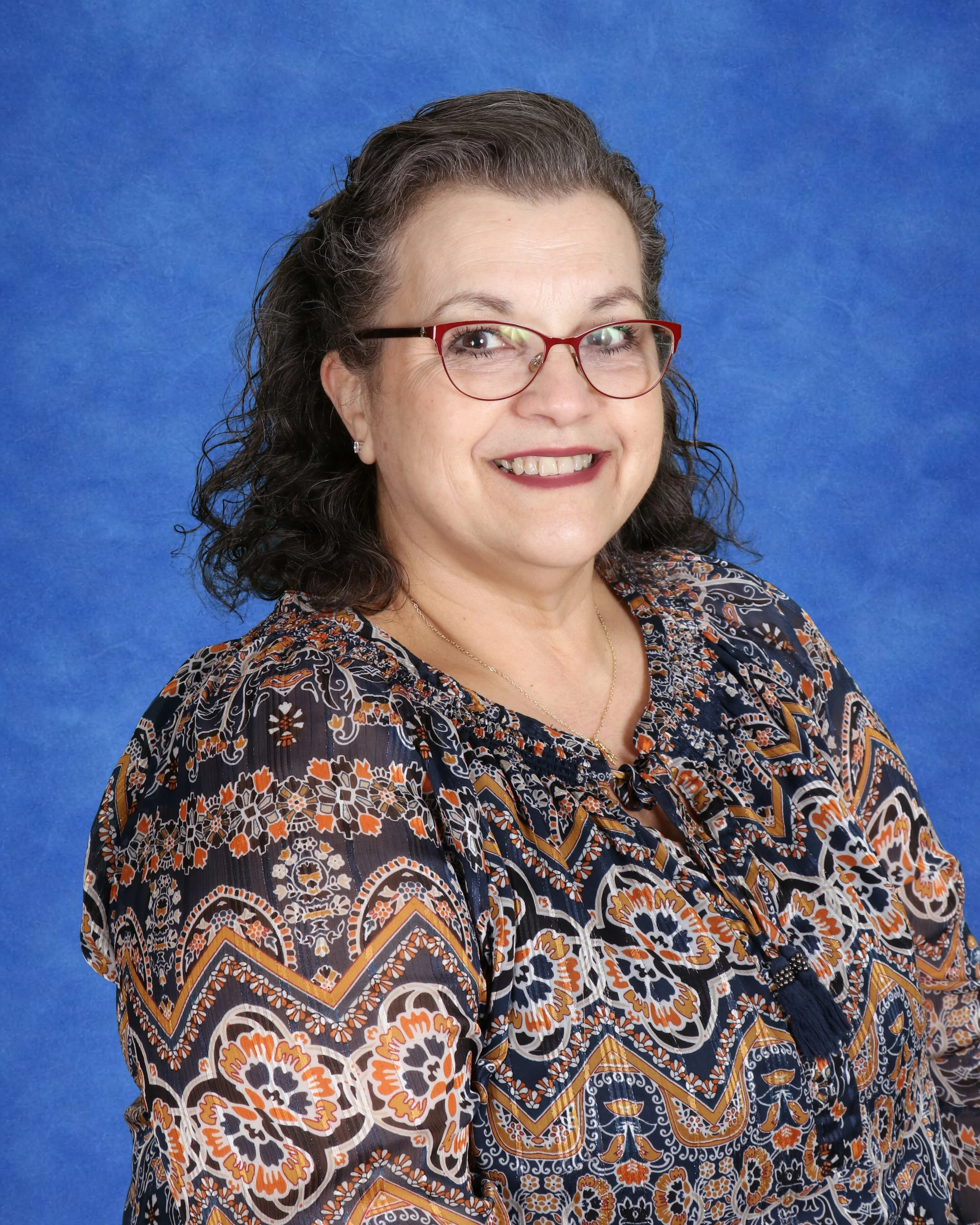 A smiling woman with glasses and long curly hair, wearing a navy blue blouse with white floral patterns, standing against a white brick wall.