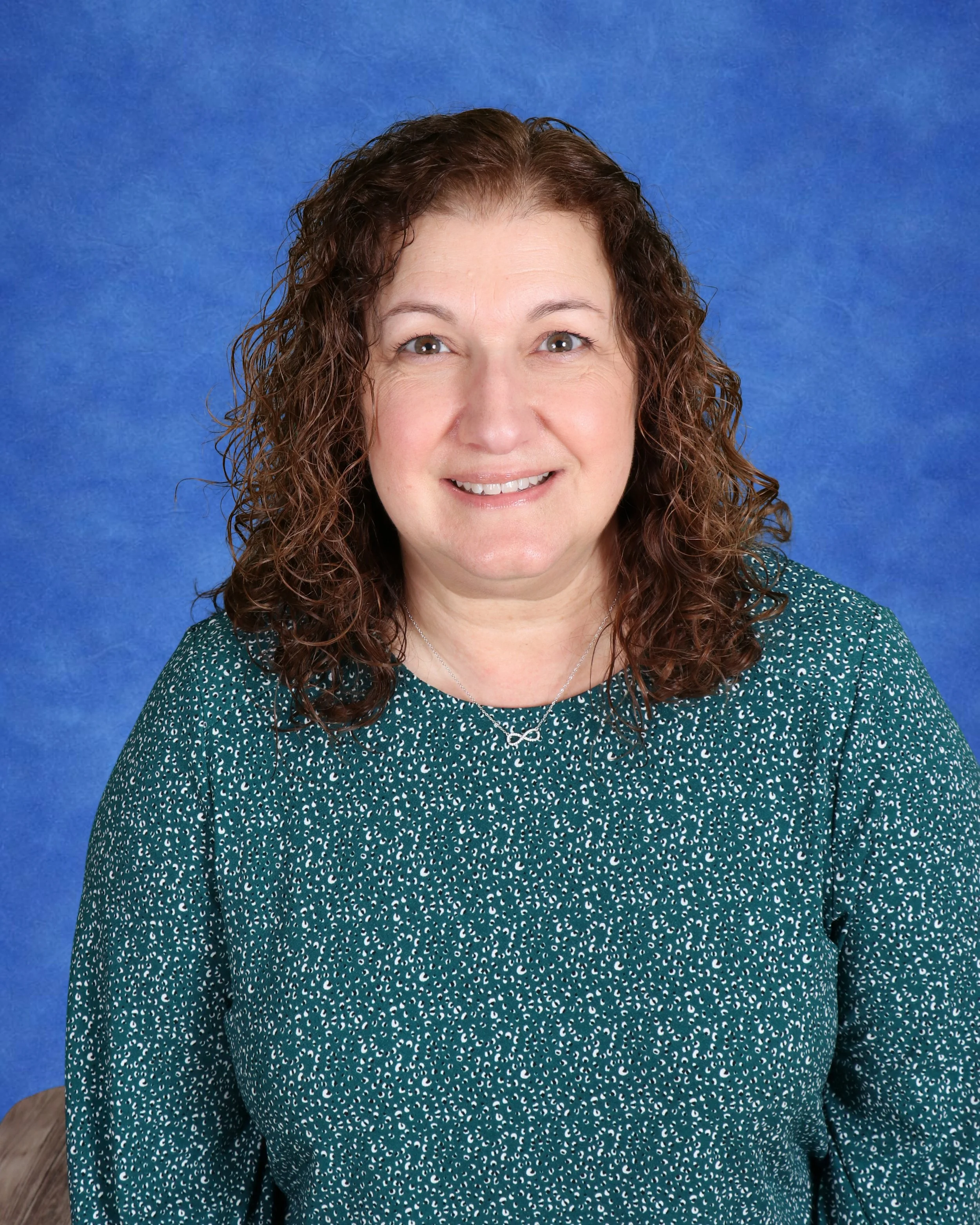Headshot of a woman with shoulder-length curly hair, wearing a denim jacket and a black top, standing against a light gray cinder block wall.