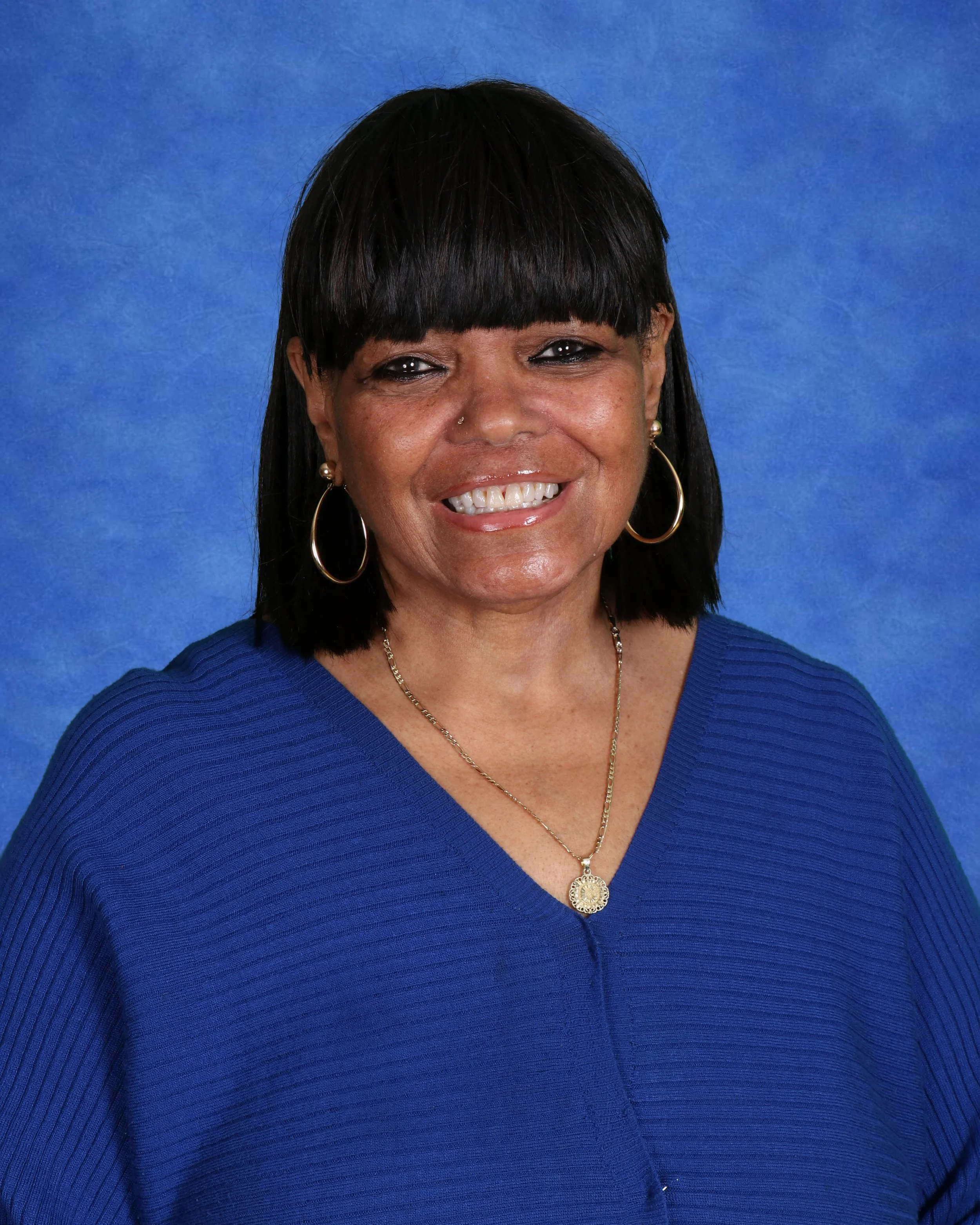 Smiling woman with earrings and necklace standing in front of a white brick wall.