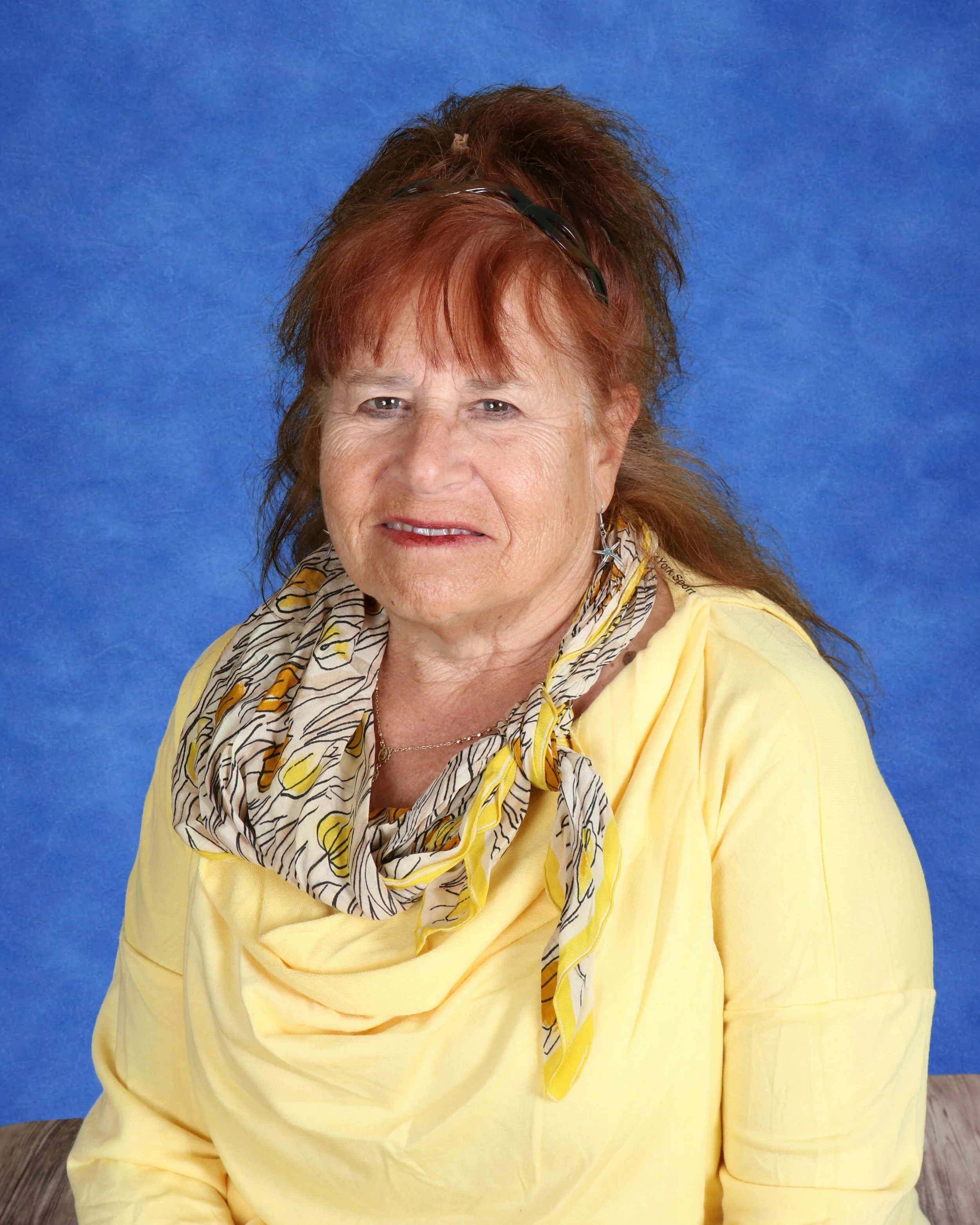 Older woman with reddish-brown hair tied in a bun with a light gray headband, smiling, wearing a blue top, standing in front of a white cinder block wall.