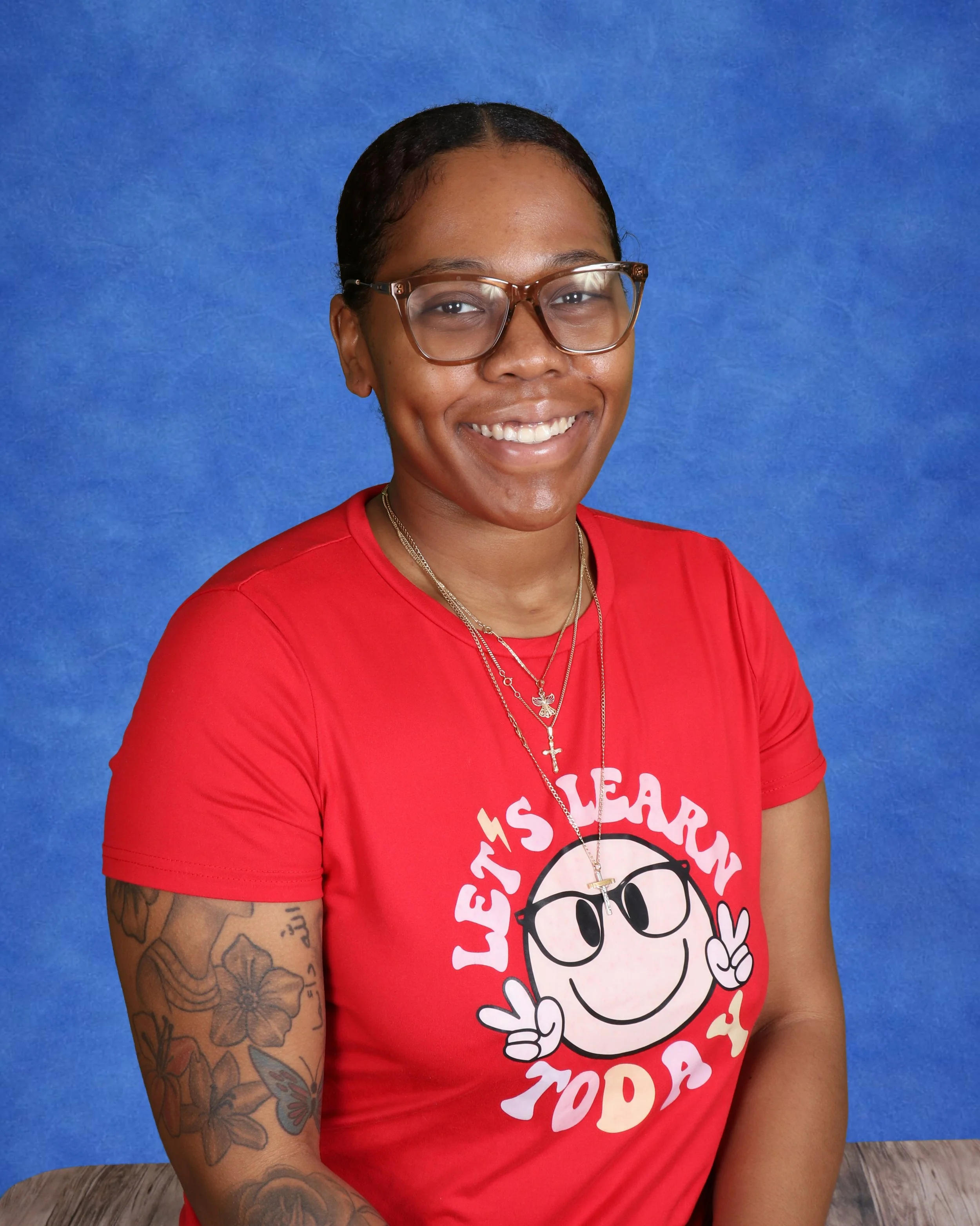 Young woman with glasses, wearing a pink headband, tie-dye t-shirt, and layered necklaces, smiling in front of a light-colored wall.