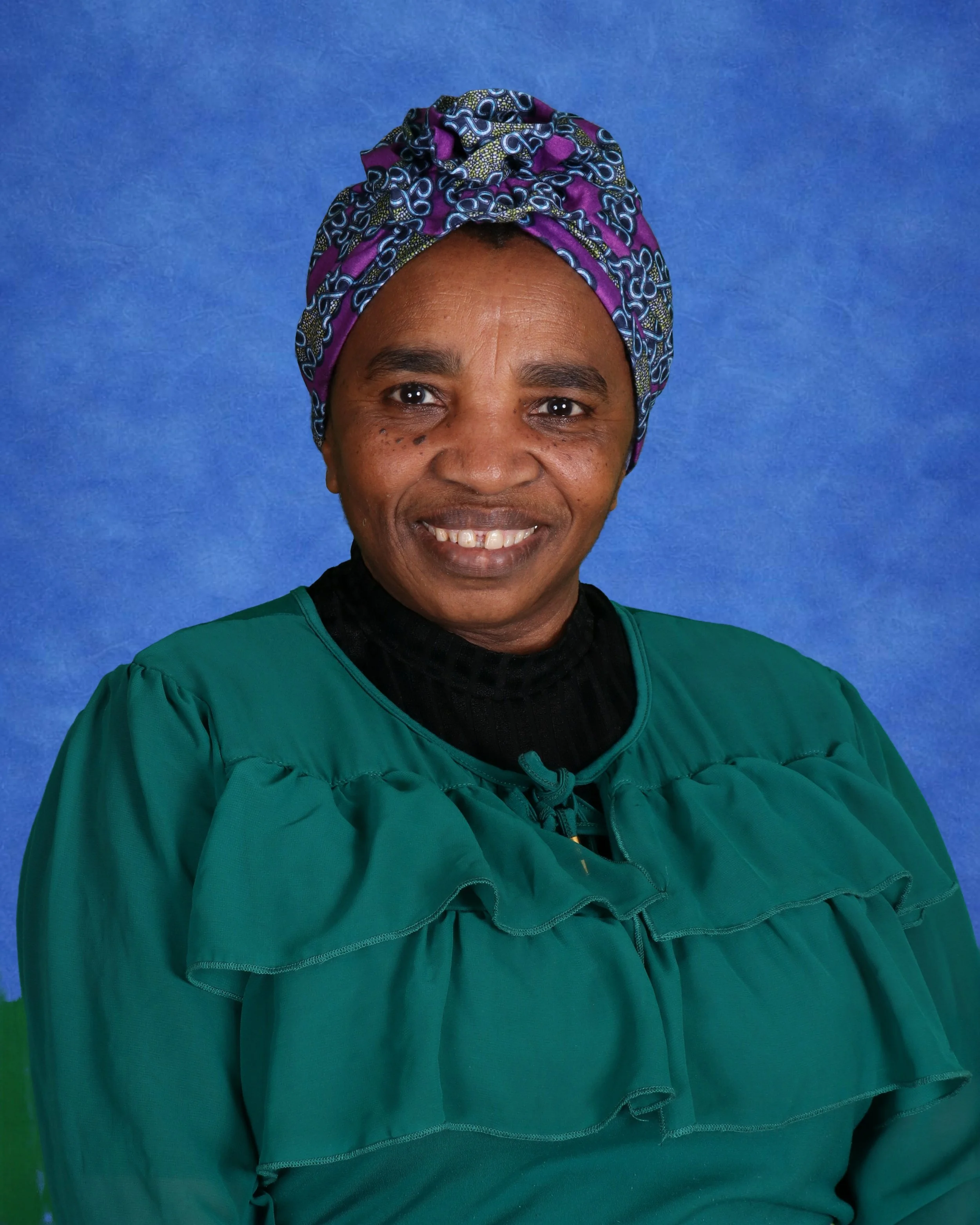 Woman wearing a colorful headwrap and a floral blouse standing against a white brick wall.