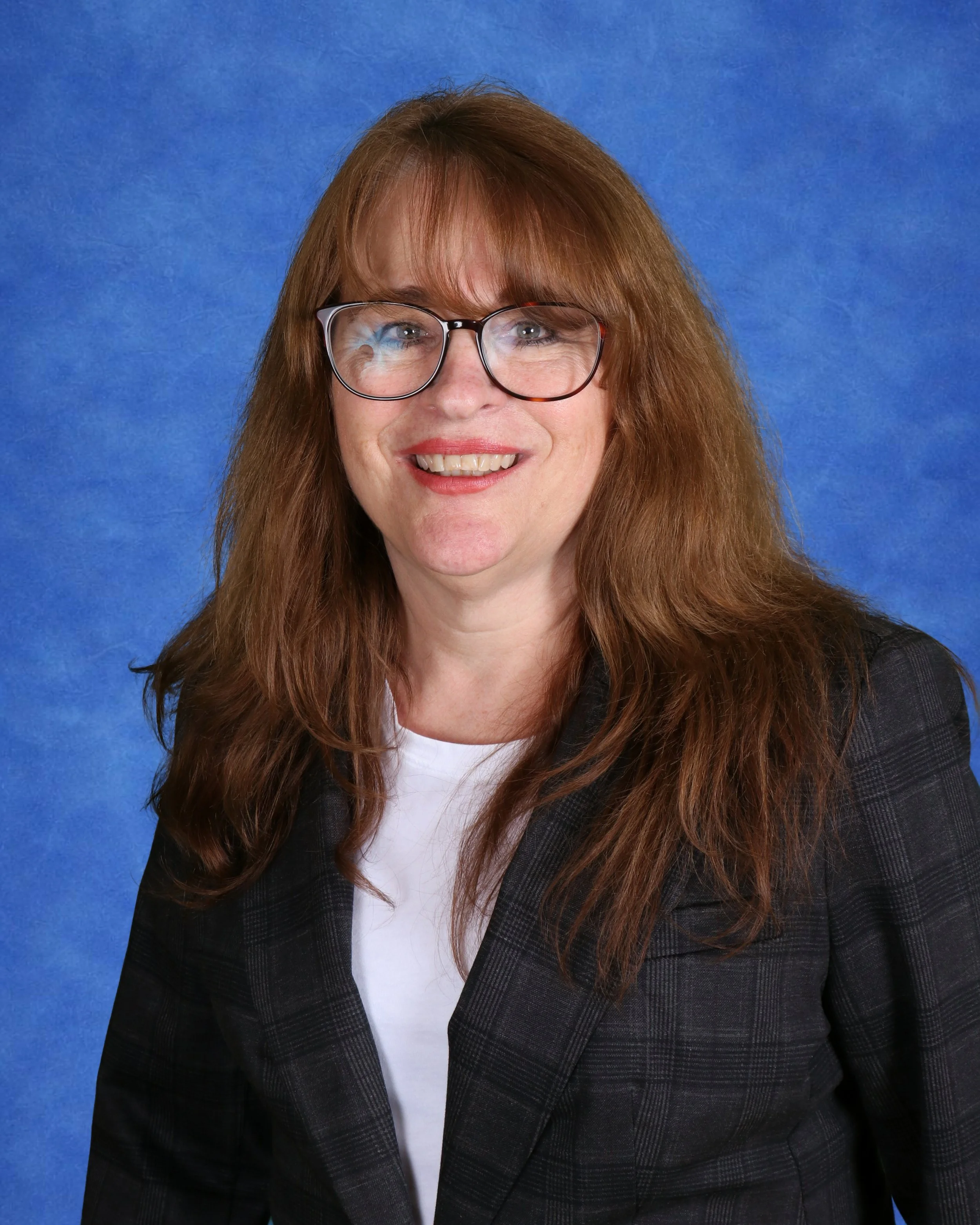 A woman with long, wavy red hair smiling in front of a light-colored cinder block wall, wearing a black turtleneck and a black-and-white checkered jacket.
