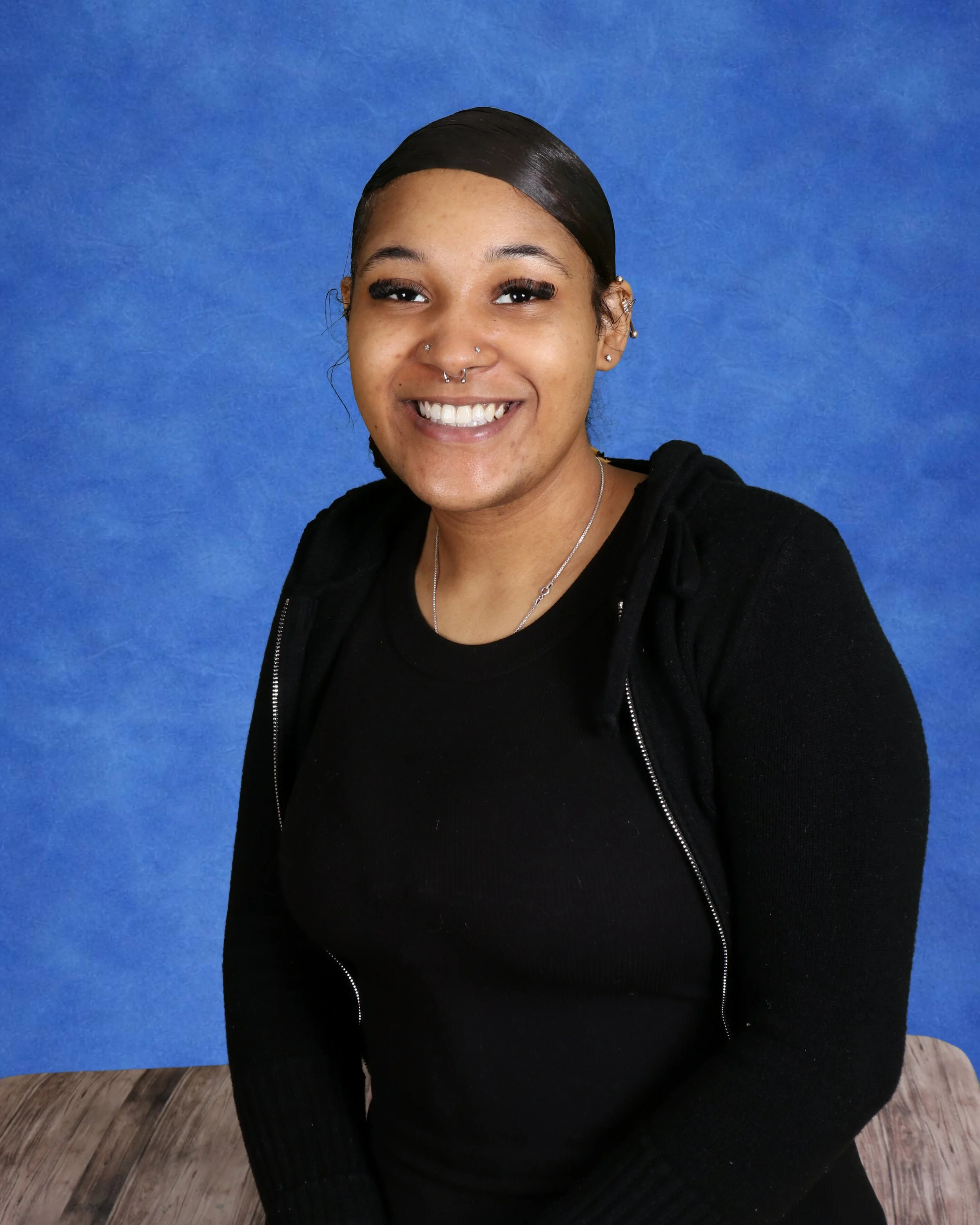 A young woman with curly hair and a septum piercing smiling in front of a light gray wall with a clock.