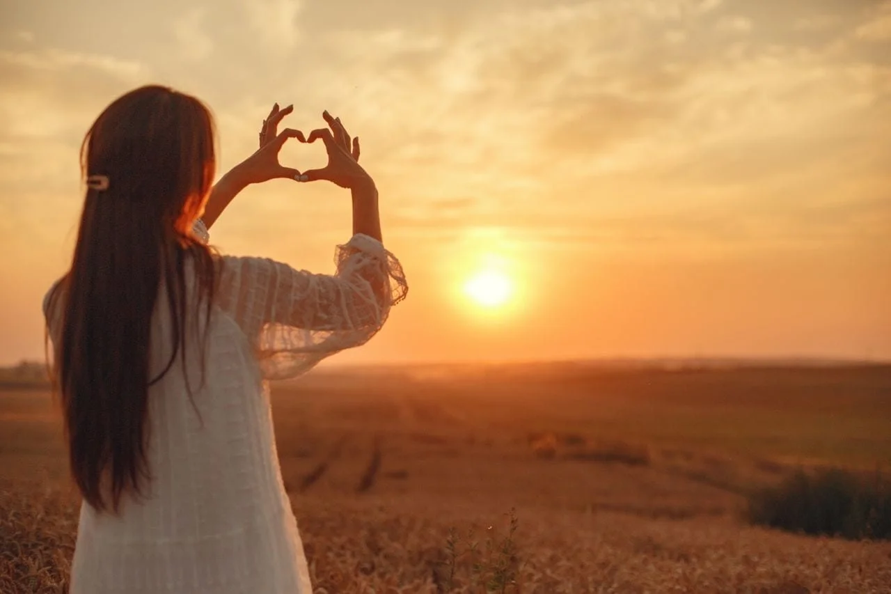 woman in white top making a heart with her fingers towards a setting sun