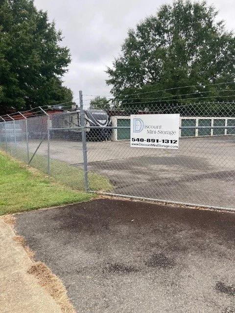 Chain-link fence surrounding a mini storage facility with a sign reading 'Discount Mini Storage' and contact information. Trees visible in the background.