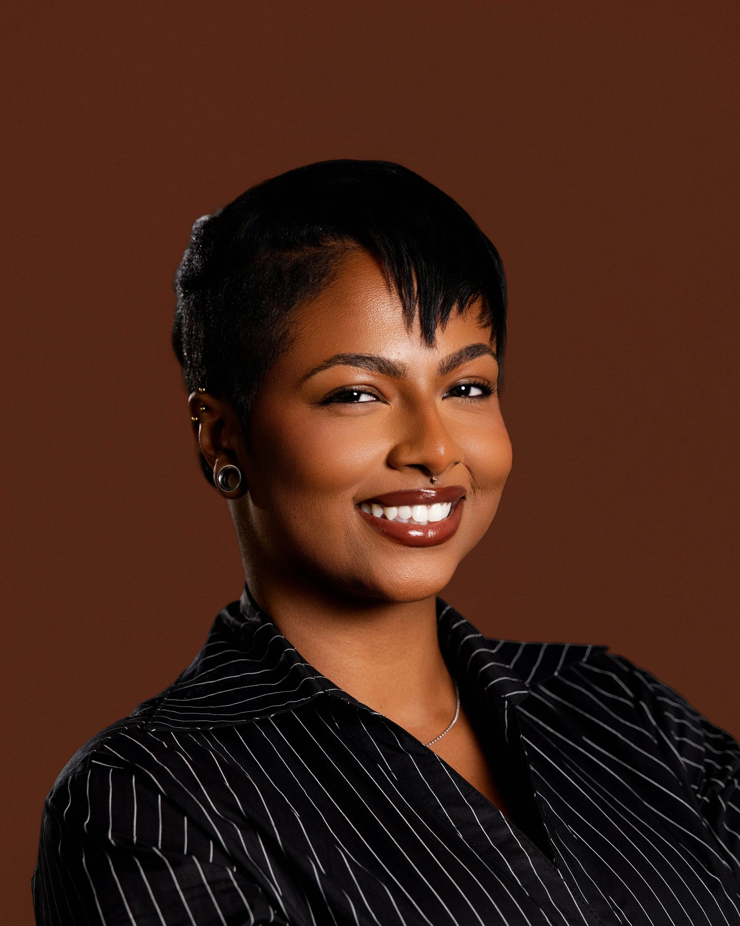 Close-up of a woman with short, black hair styled in a side part, smiling, wearing earrings, a necklace, and a black shirt with white stripes, against a brown background.