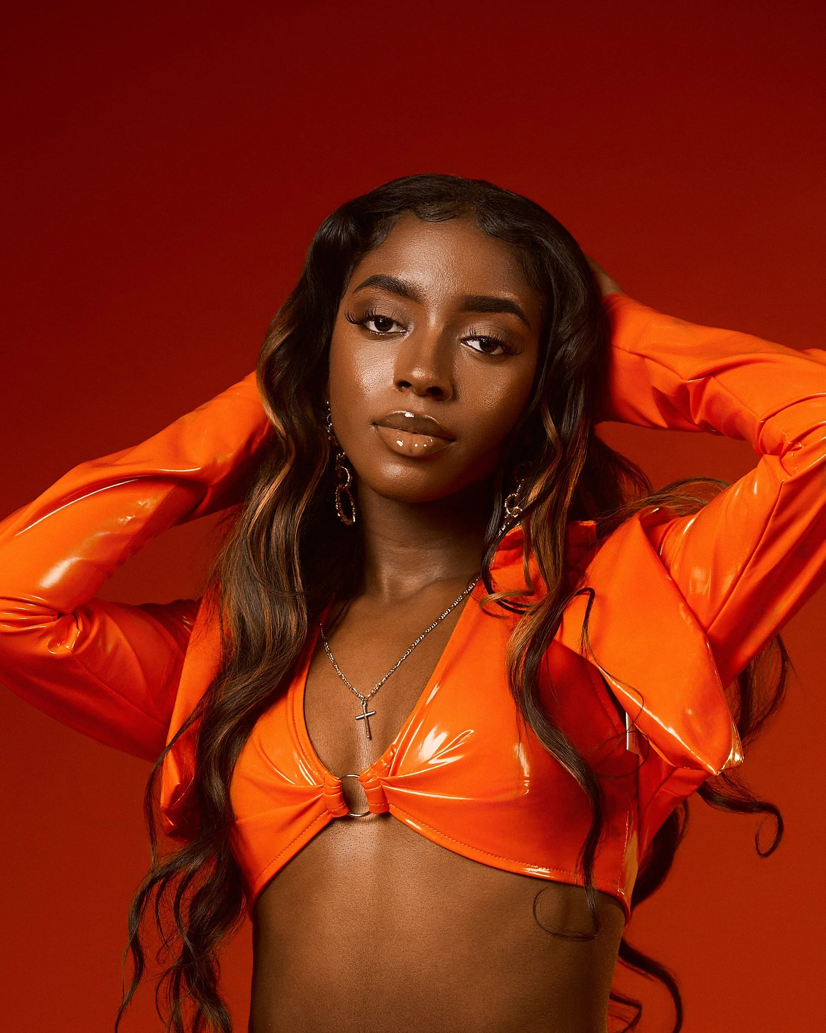 A woman with long wavy hair wearing an orange shiny top with a deep neckline, a cross necklace, and gold earrings, posing with her hand behind her head against a red background.