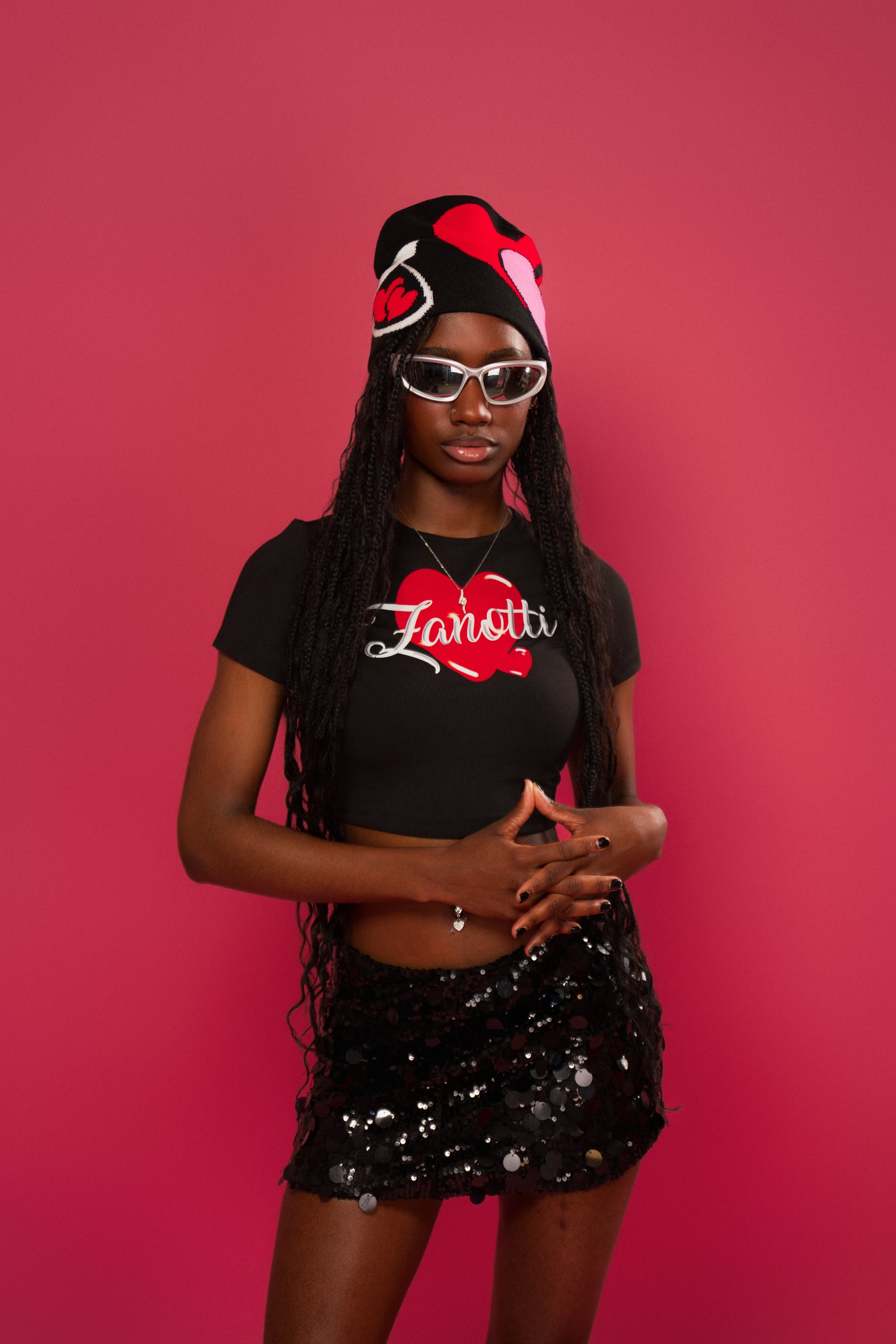 A young woman wearing a black T-shirt with red and white text, a beanie with red, pink, and black colors, white sunglasses, and a black sequined skirt, posing against a pink background.