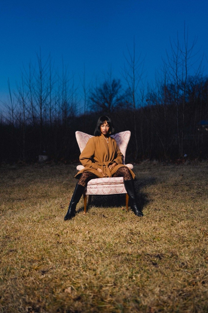 A woman sitting on a pink armchair outdoors at dusk, wearing a tan coat, animal print leggings, and black boots, with a natural background of leafless trees and a clear dark blue sky.