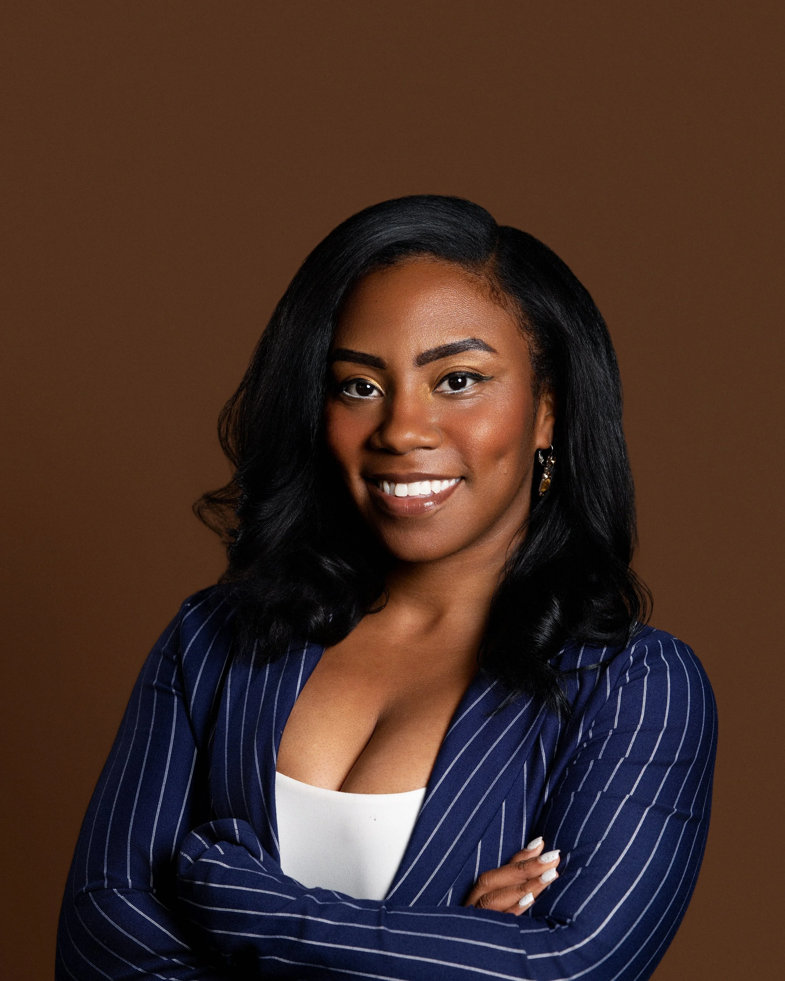 Close-up of a woman smiling, with long black hair styled with waves, wearing a navy blue pinstripe blazer over a white top, standing against a brown background.
