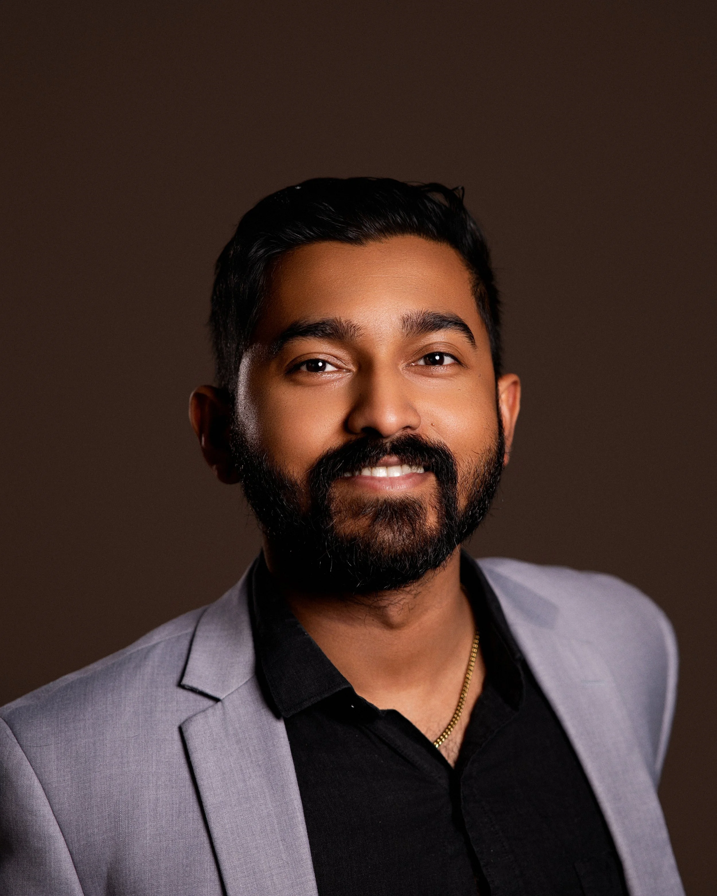 Close-up of a smiling man with dark hair, beard, and mustache, wearing a gray blazer, black shirt, and gold chain, against a dark background.