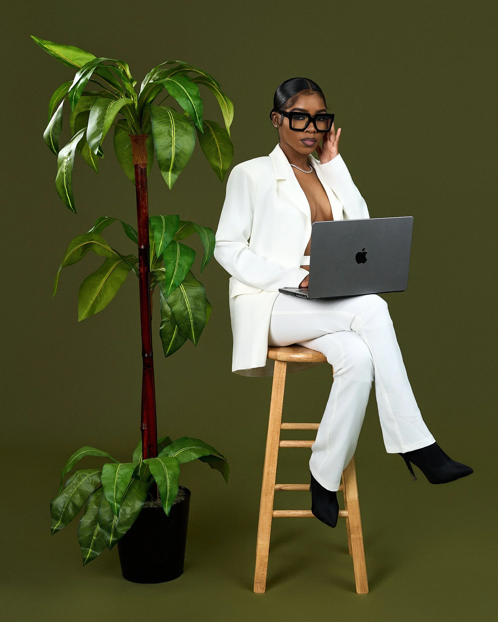 A woman dressed in a white suit with black shoes sitting on a wooden stool, adjusting her black glasses, next to a large potted green houseplant in front of a plain olive green background.