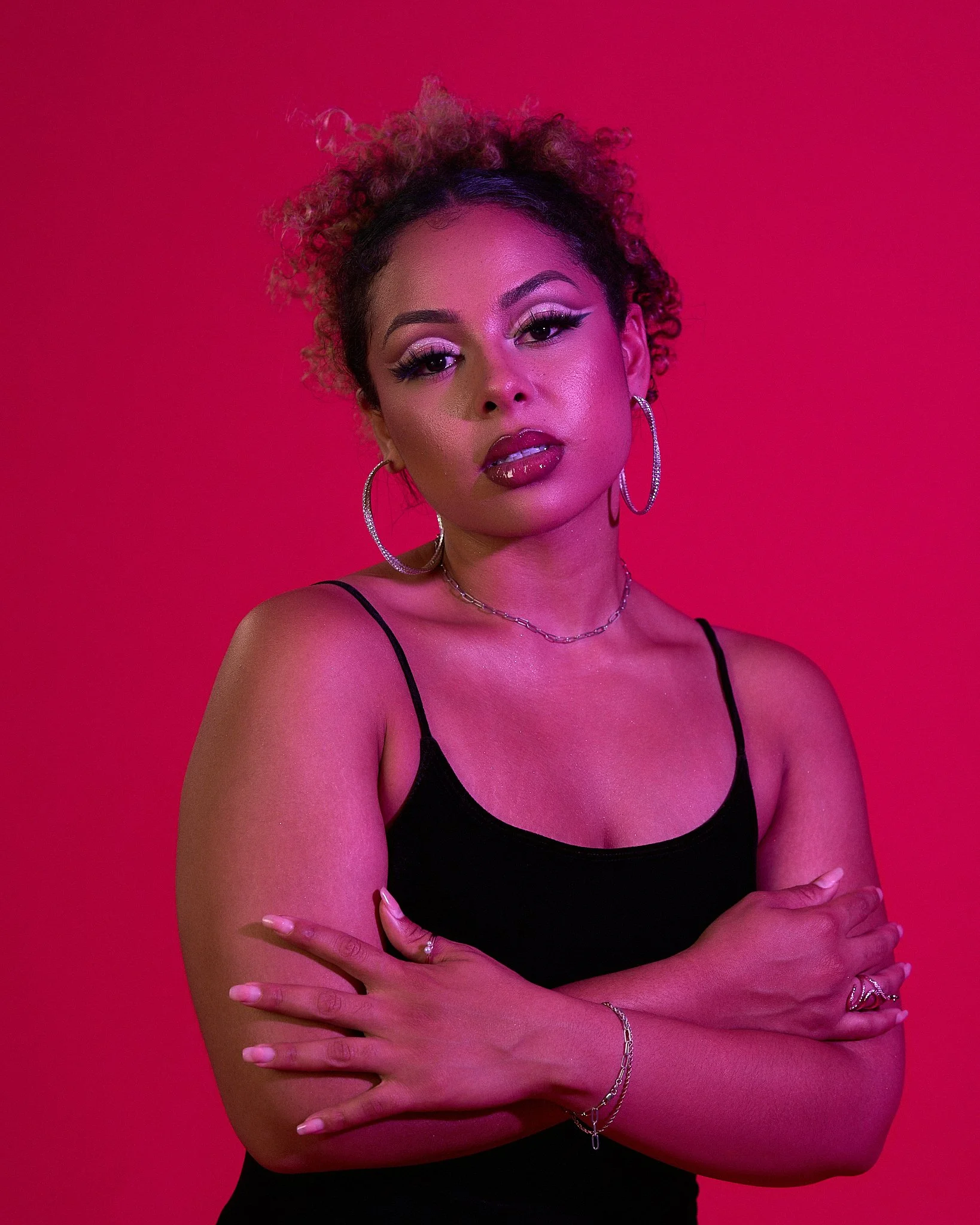 A woman with curly hair in front of a pink background, wearing a black top and hoop earrings, with crossed arms and jewelry.