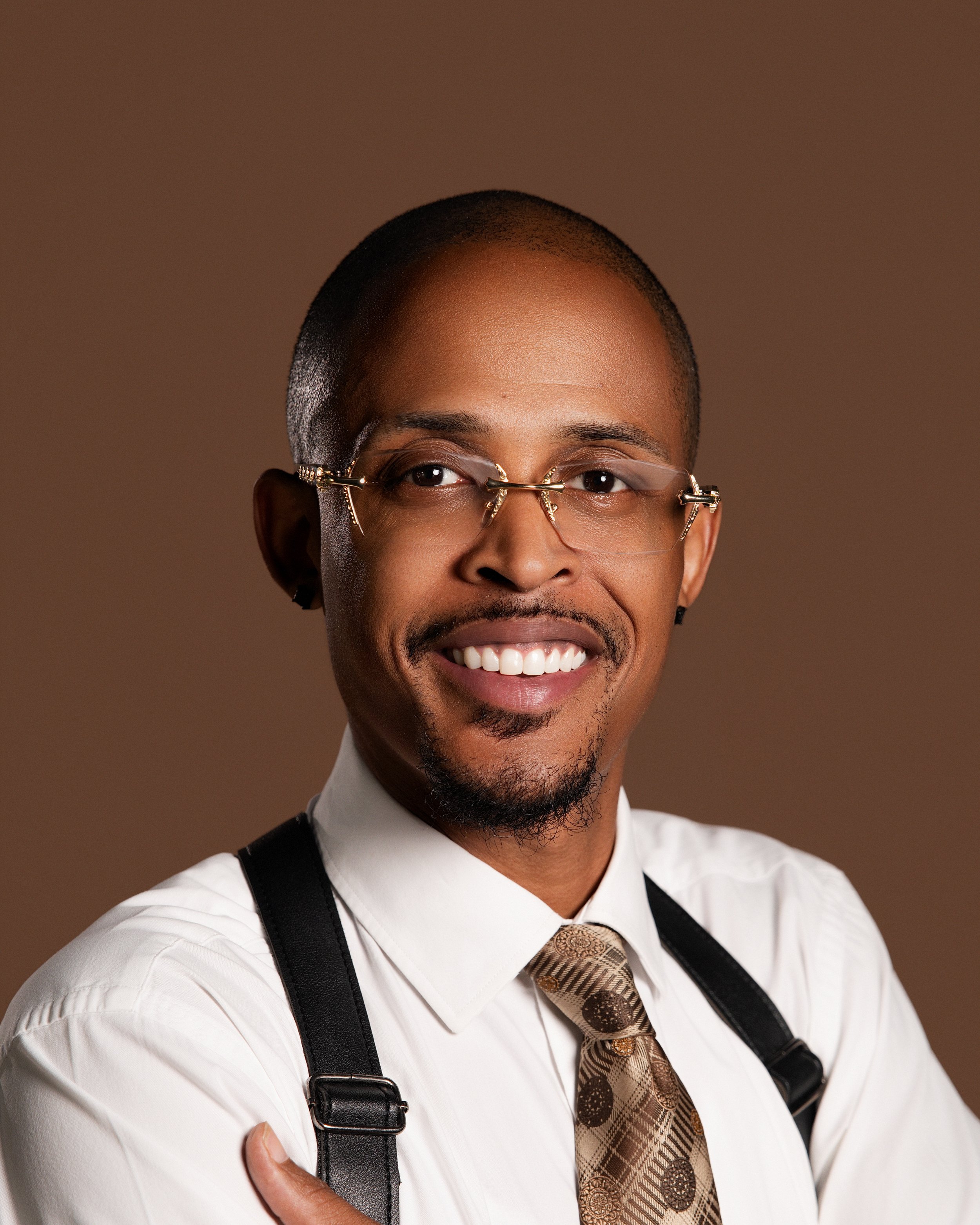 Close-up of a smiling African American man wearing glasses, a white shirt, a patterned tie, and suspenders against a brown background.