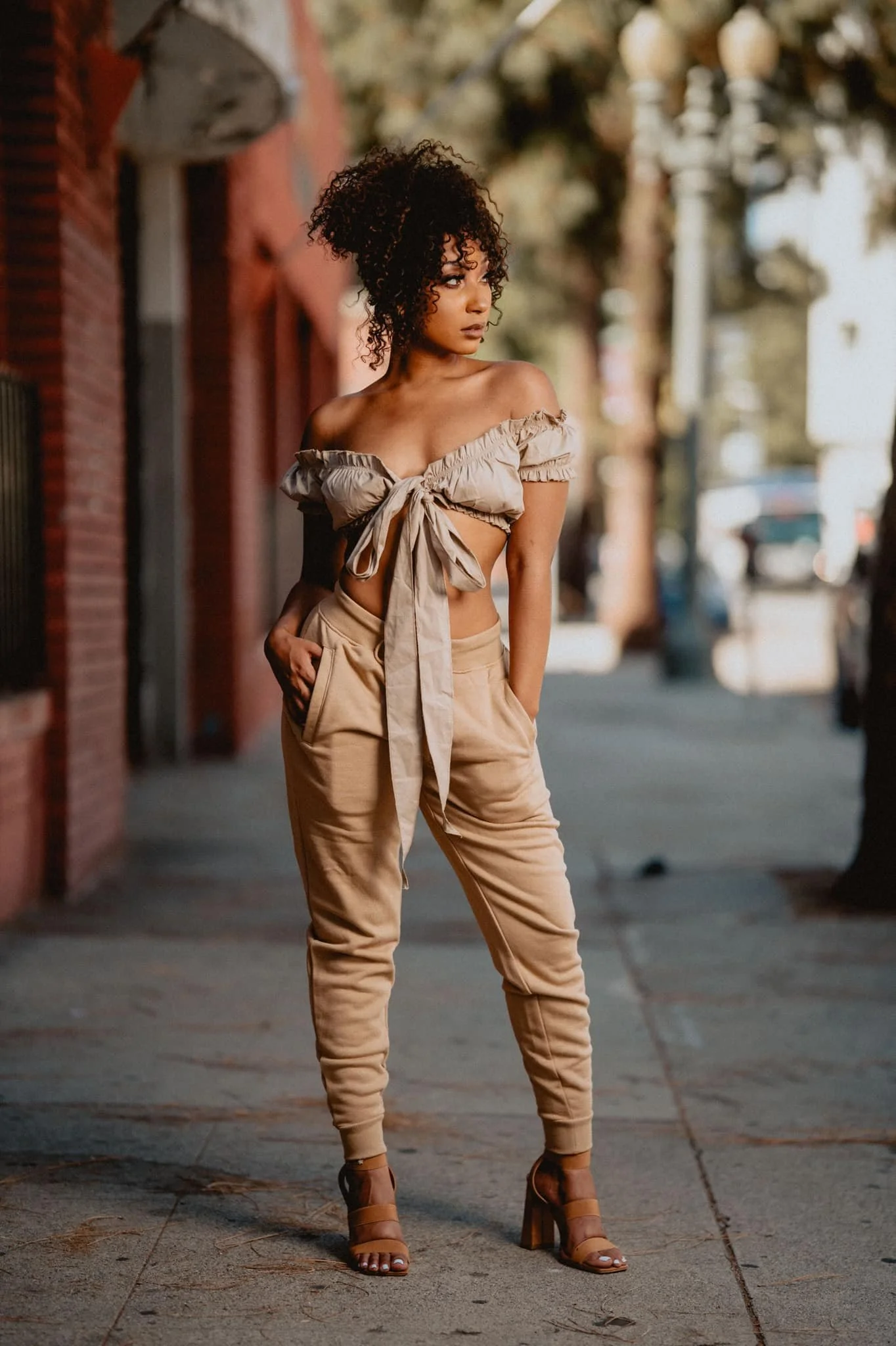 A woman with curly hair standing on a city sidewalk during daytime, wearing beige off-shoulder top with a front tie and high-waisted beige pants, heels, with a blurred background of trees, buildings, and parked cars.