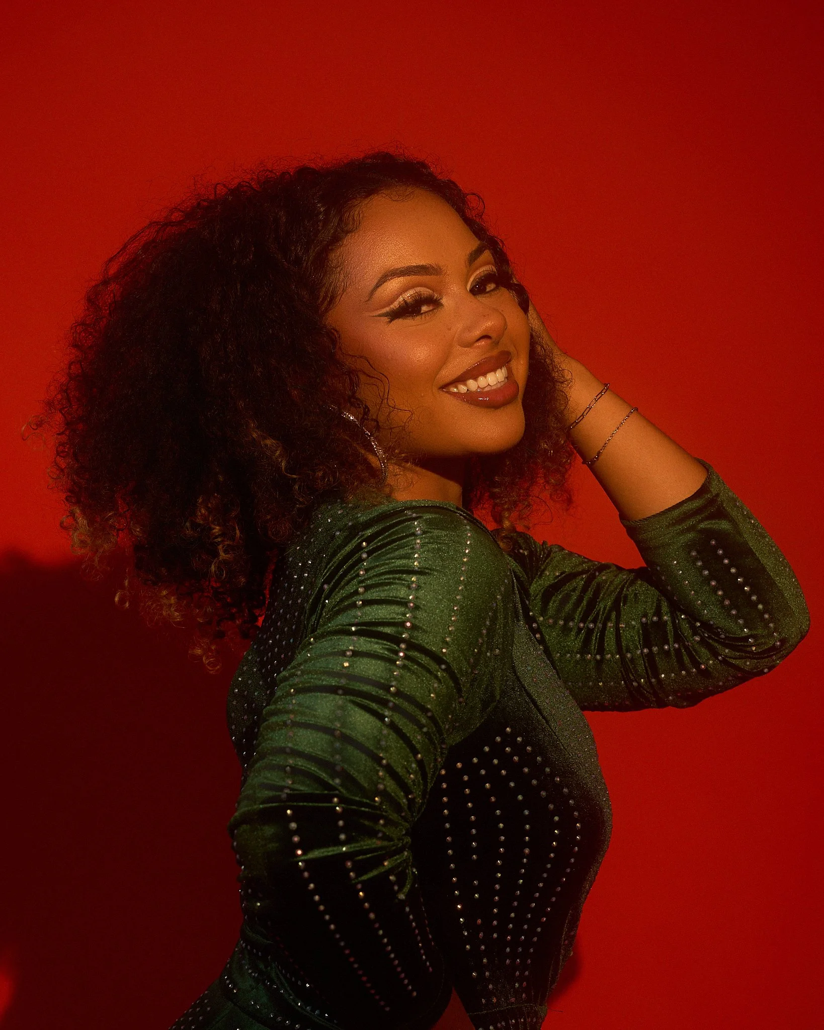 A woman with curly hair, wearing makeup, a green, shiny, studded top, and jewelry, smiling against a red background.