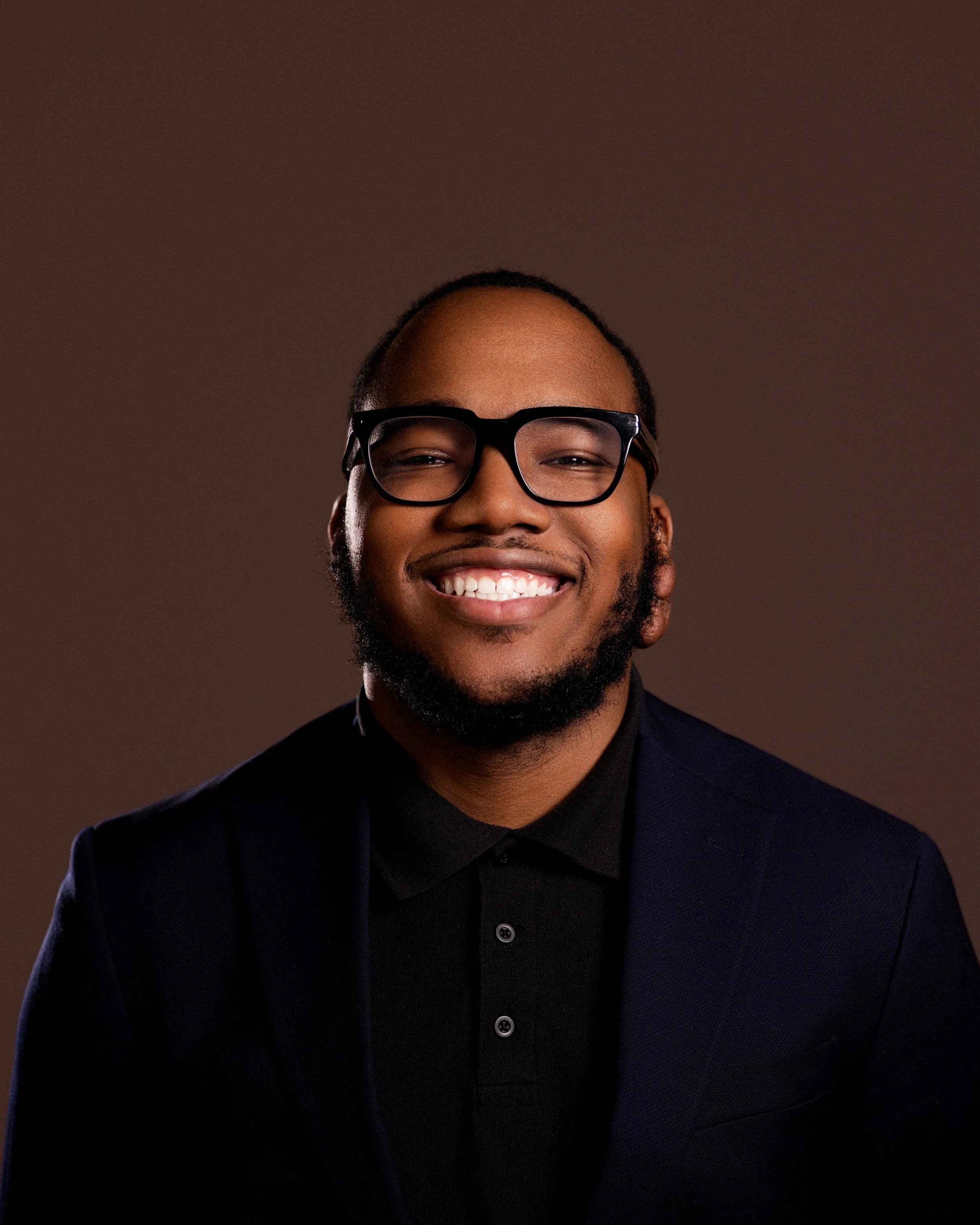 Smiling man with glasses wearing a dark suit and black shirt, portrait against a dark background.