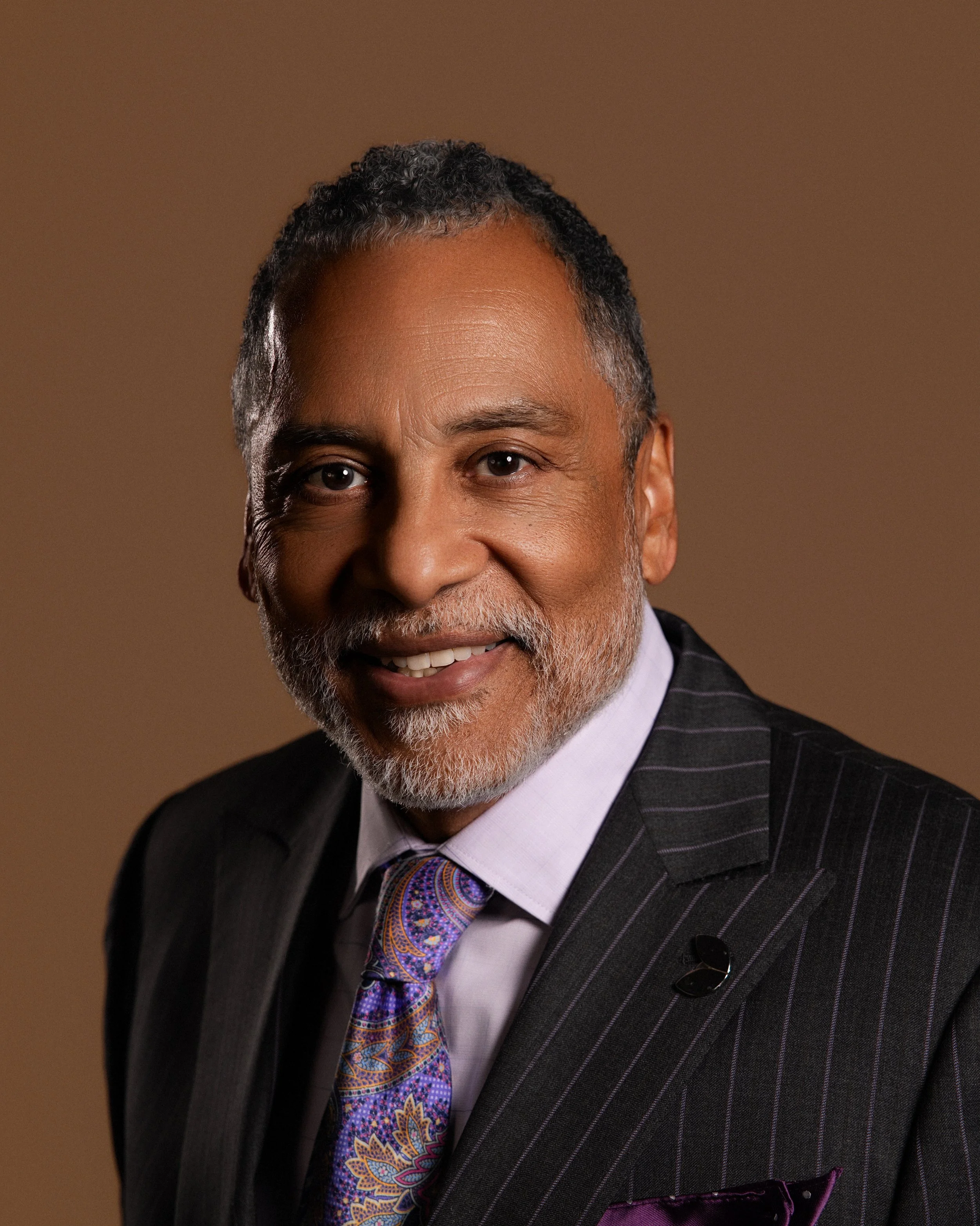 Close-up portrait of a smiling African American man with gray hair and beard, wearing a gray suit with pinstripes, a patterned purple tie, white shirt, and purple pocket square, against a brown background.