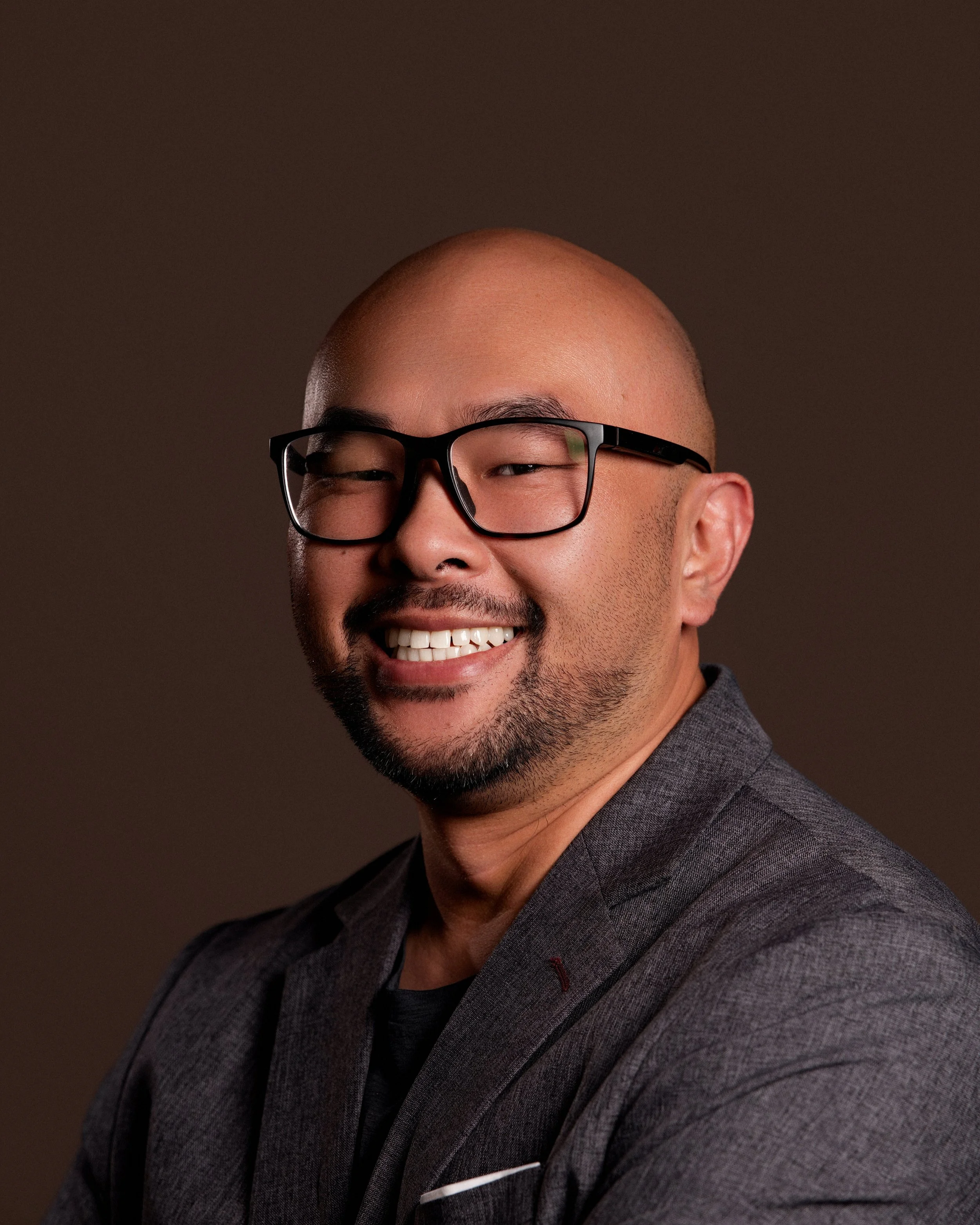 Close-up of a smiling man with a bald head, wearing black-rimmed glasses, a gray blazer, and a dark shirt, against a brown background.