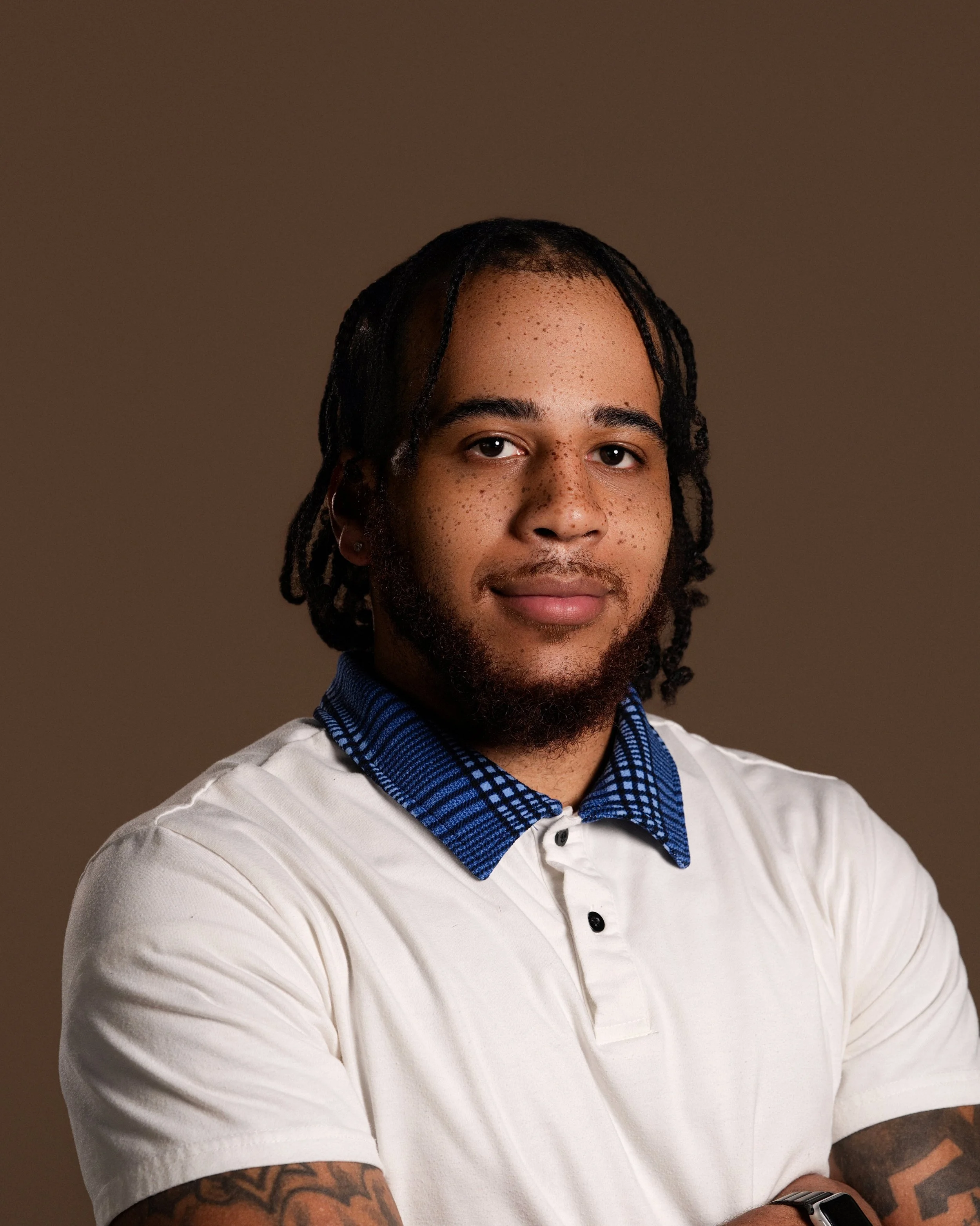 Close-up of a young man with braided hair, freckles, and a beard, wearing a white shirt with a blue patterned collar, standing against a plain dark background.