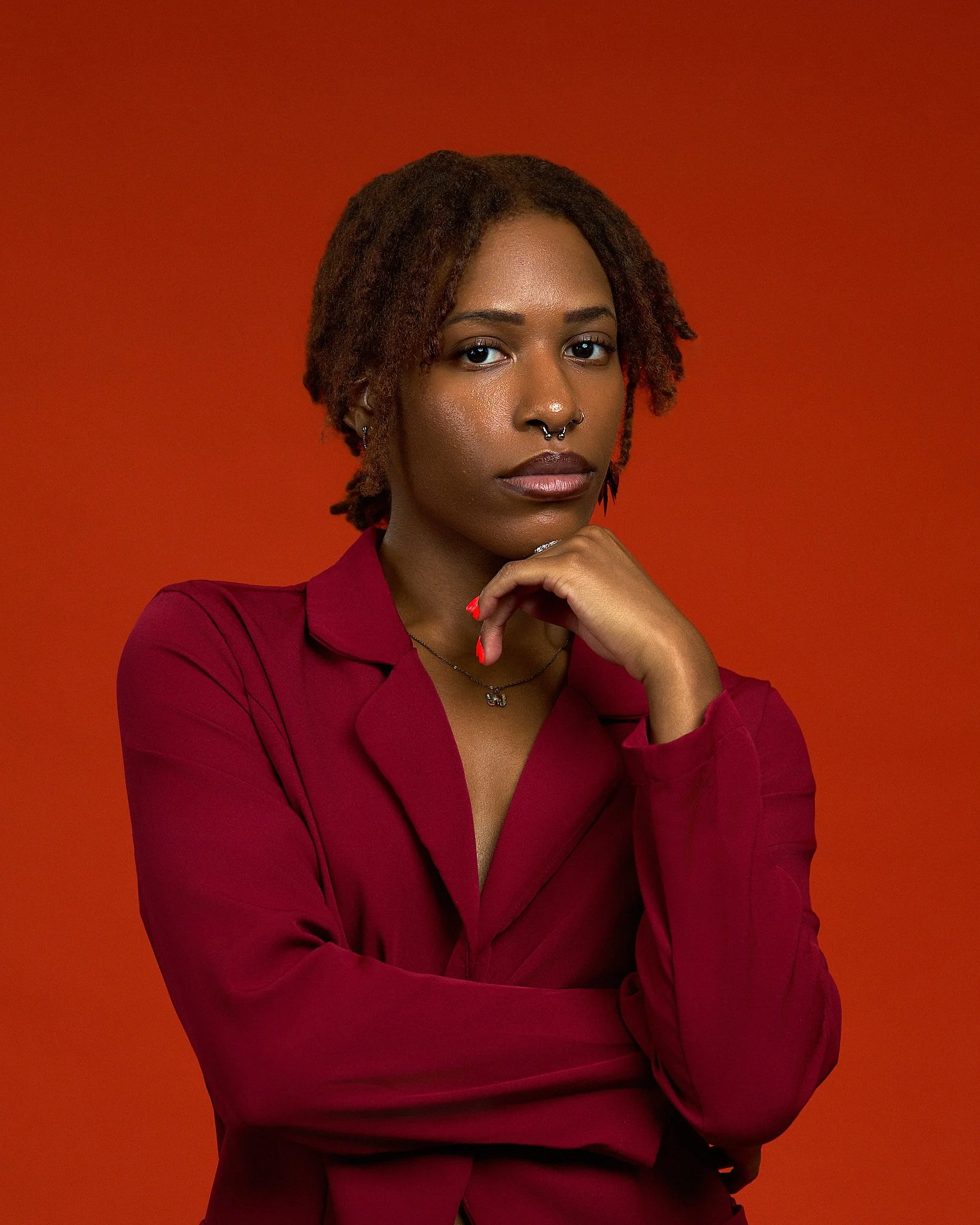 A woman with dark skin and reddish-brown dreadlocks, wearing a burgundy top, with her right hand resting under her chin, against an orange background.