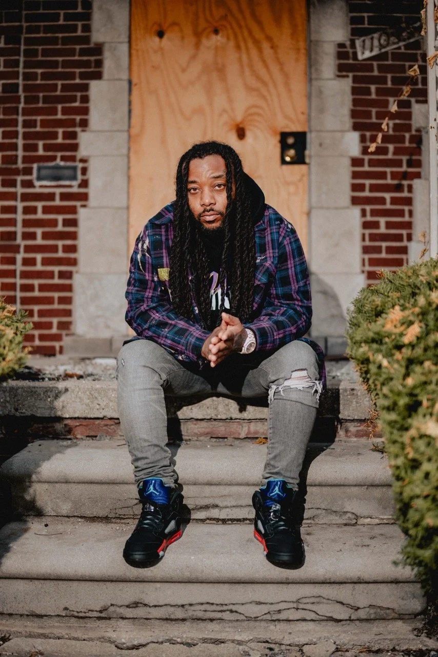 A man with long dreadlocks sitting on front steps of a building with brick and stone walls, wearing a purple plaid jacket, gray ripped jeans, and black sneakers with red and blue accents.