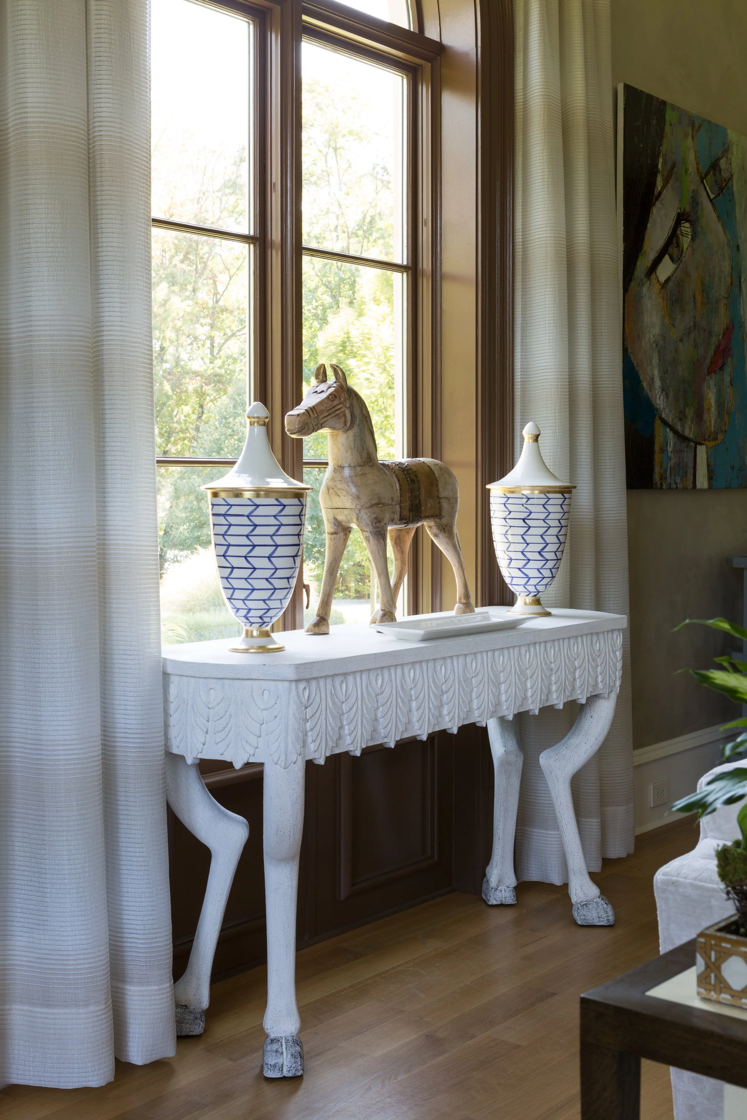A white wooden console table with carved leaf details and horse leg-shaped supports, decorated with a wooden horse figurine and two blue and white ceramic vases with lids, positioned in front of a large window with white curtains, in a room with a painting on the wall and hardwood floor.