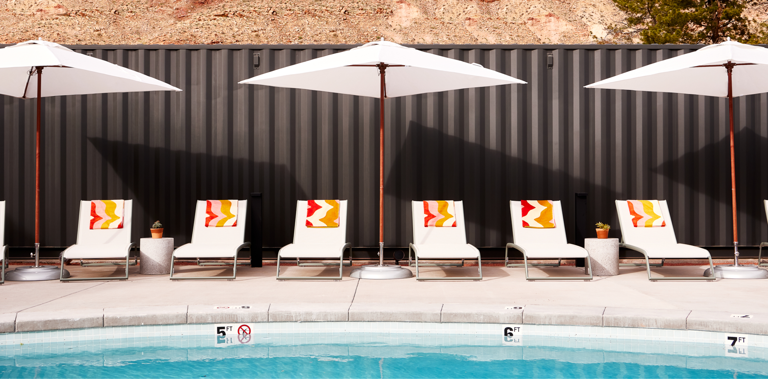 A row of sun bathing chairs sit against a pool in the Moab desert at Field Station with red and orange towels under umbrellas.