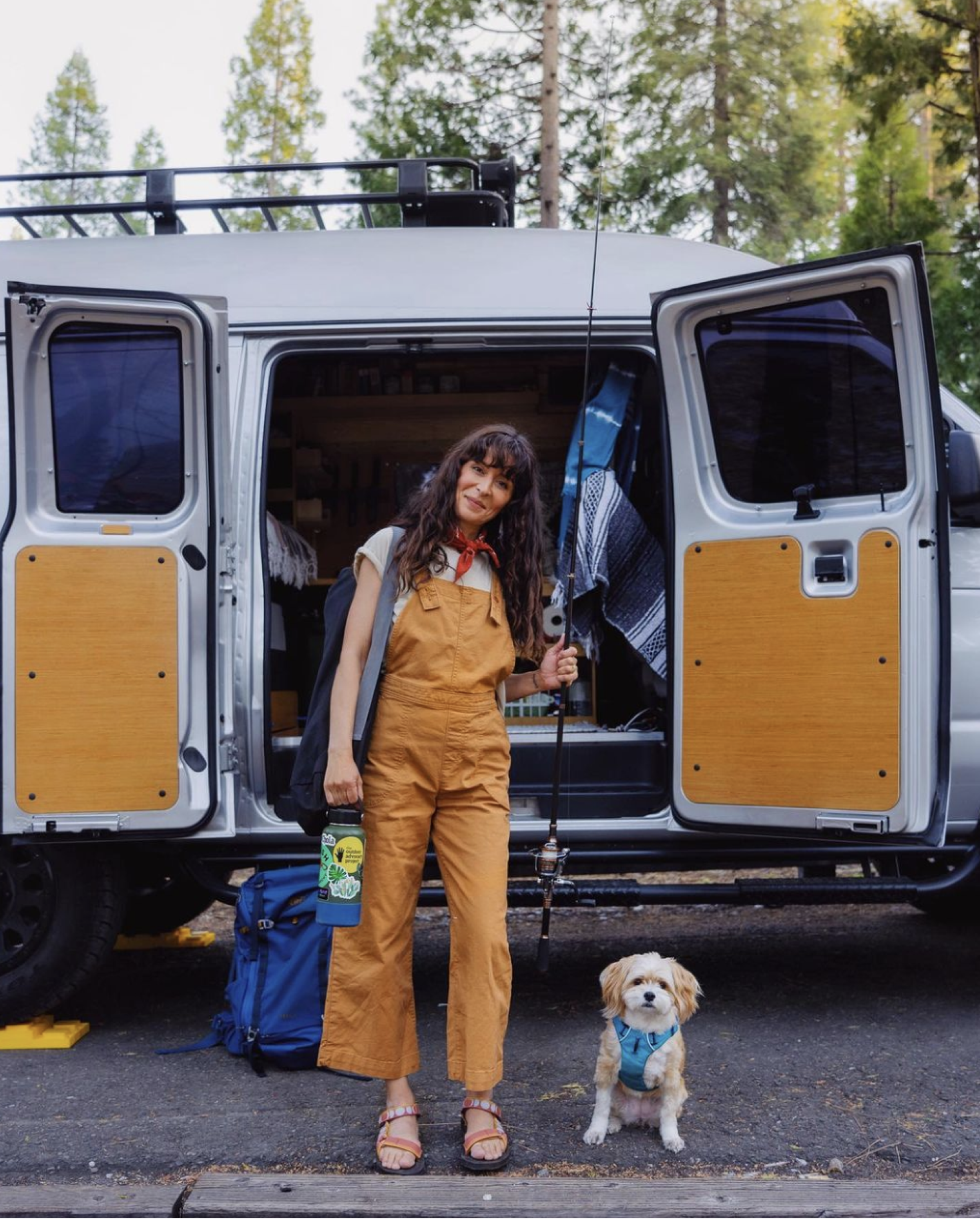 A women with dark brown hair, brown overalls, and a red bandana around her neck stands in front of a camper van holding a fishing rod. A little white dog stands next to her with a curious look on her face.