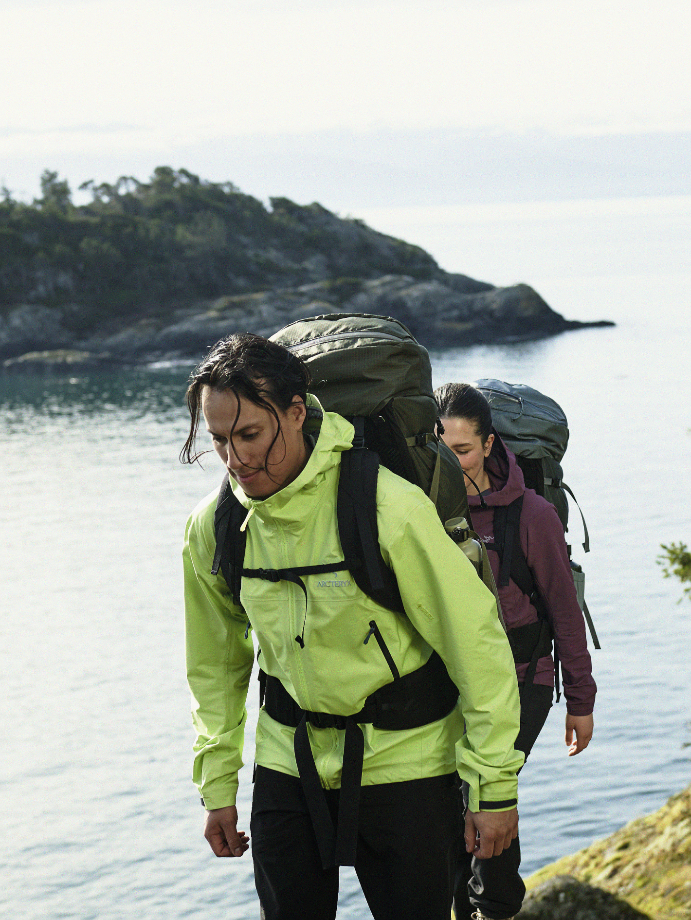 In front of a coastal scene, two hikers are seen trekking. The man in front is wearing a neon green Arc'teryx Beta Jacket with a dark green backpacking pack. The woman behind him is wearing a purple Arc'teryx jacket with a backpacking pack.
