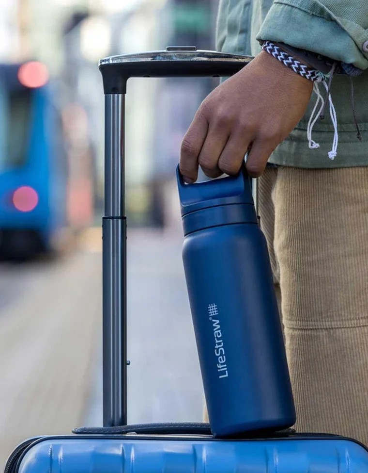 A blue LifeStraw insulated tumbler with a straw sits on a wooden table in the foreground. In the background, a woman in light purple clothing is seated in a cross-legged meditation pose on a mat surrounded by potted plants on a wooden deck.