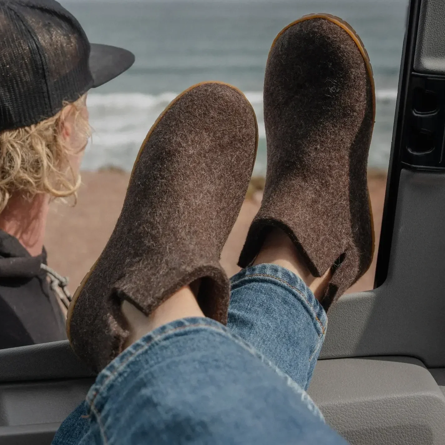 Two people wearing felt Glerups slippers, sitting on a rock by the beach, with their feet up, overlooking ocean waves.