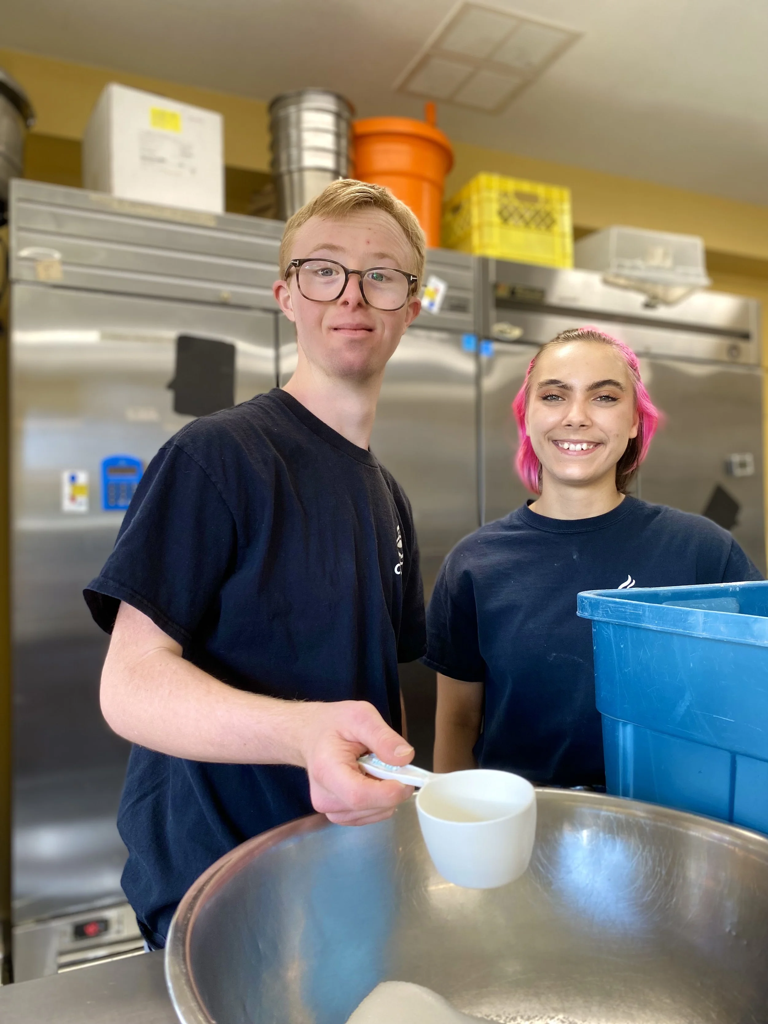 Two young people standing in a commercial kitchen, smiling at the camera; one holding a measuring cup over a large mixing bowl, with kitchen shelves and equipment in the background.